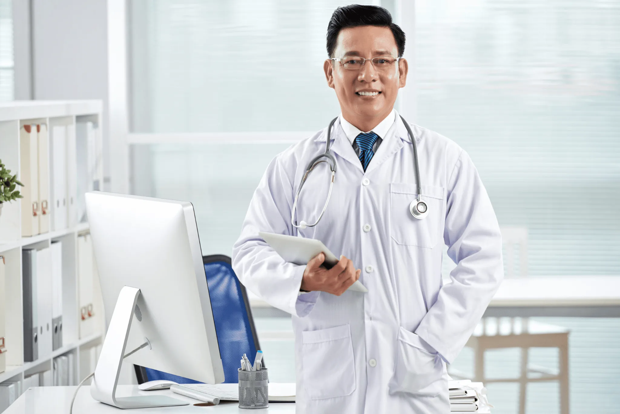 Asian male physician wearing a white coat with a stethoscope around his neck, left hand on his coat front pocket, right hand holding a tablet