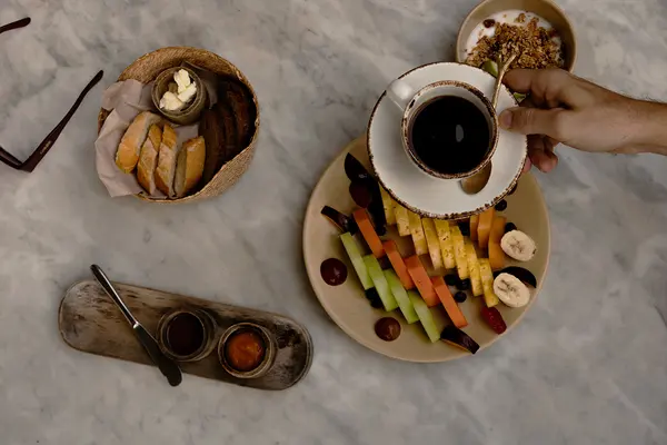 Breakfast dishes served at Sky Terrace, Be Tulum, including buttered bread, milk with cereal and oatmeal, fruit platter with papaya, banana, kiwi, pineapple, and a coffee cup held by a man's hand.