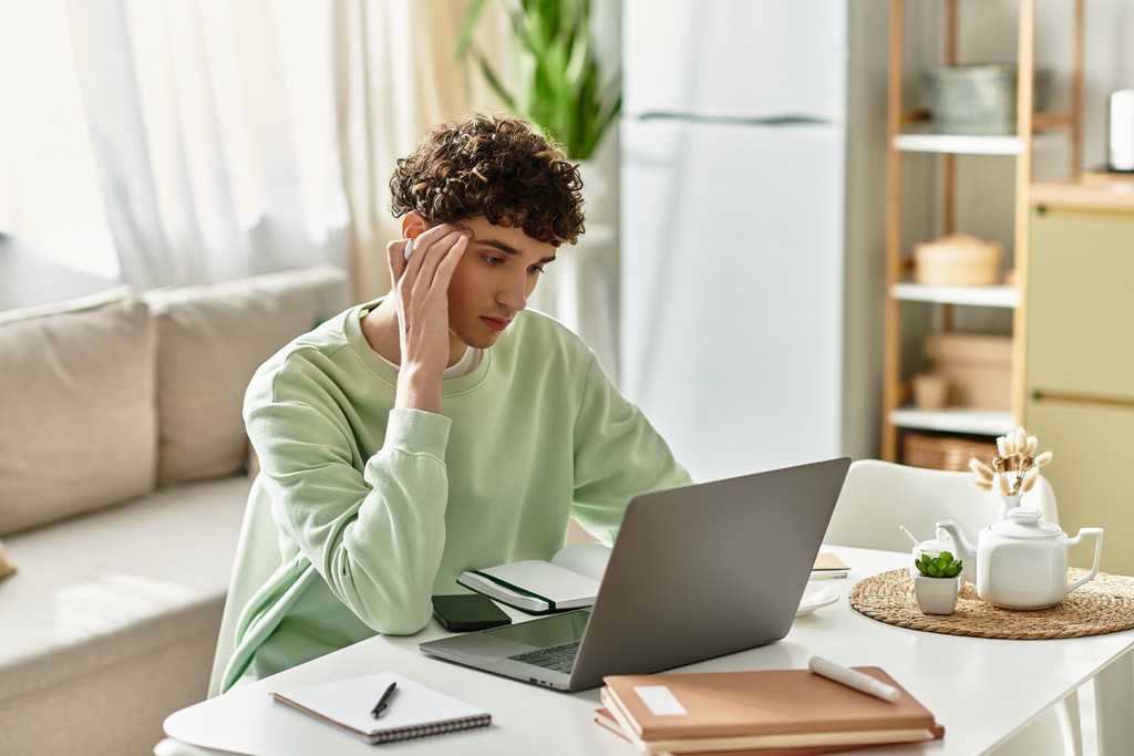 A young man in a light green sweatshirt sits at a home desk, resting his head on his hand while looking at his laptop, with a notebook and teapot nearby.