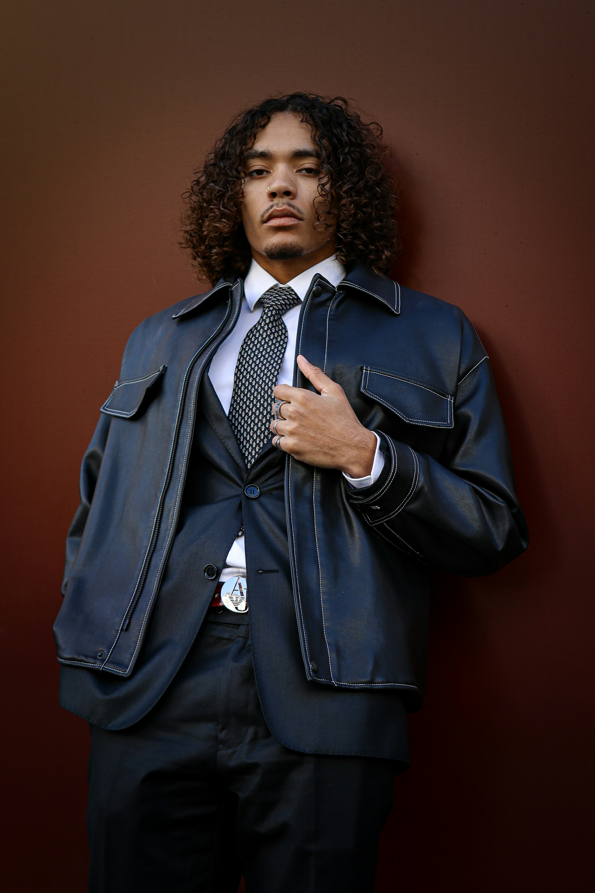 A man with curly shoulder-length hair poses against a solid dark brown background. He is wearing a formal black suit with a white shirt and patterned tie, layered under an oversized black leather jacket featuring white stitching. He has several rings on his fingers and an ornamental belt buckle.