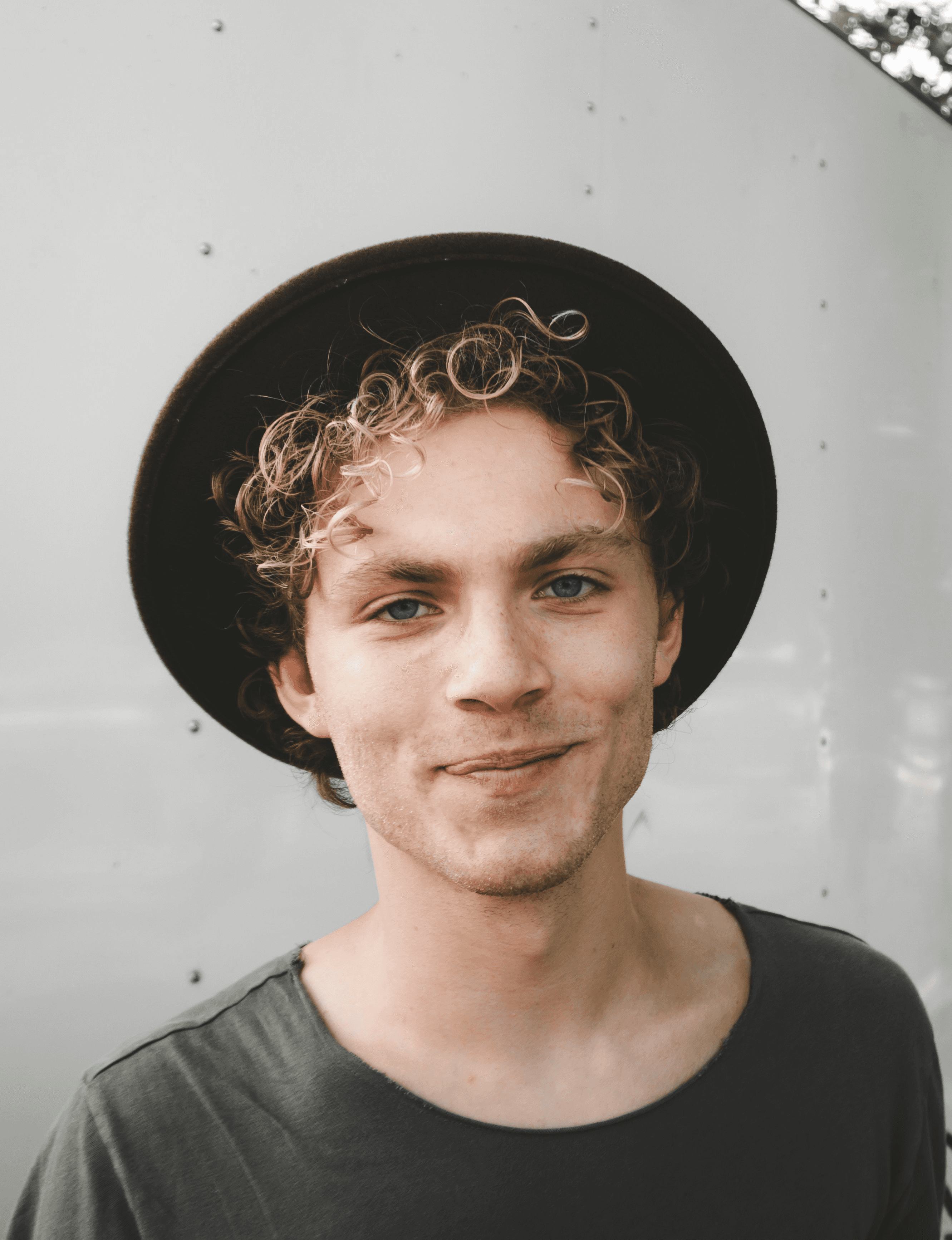 Person with curly hair in wide-brimmed hat and dark shirt standing against light background