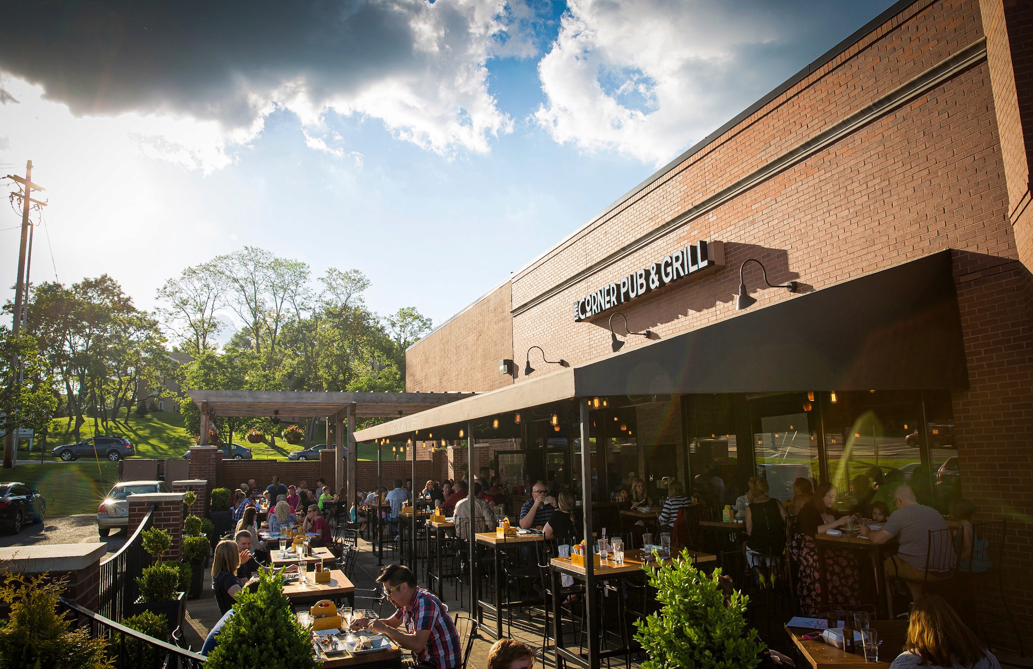 A patio scene with outdoor seating, lush greenery, and a building under a partly cloudy sky.