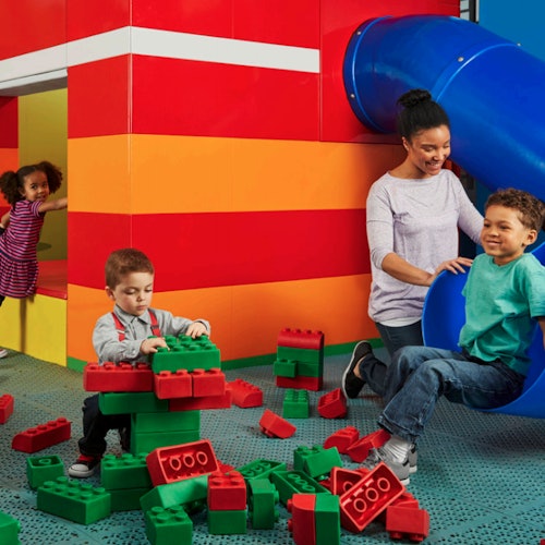 Barn leker med stora klossar nära en färgglad struktur med en blå rutschkana. En vuxen hjälper ett barn vid slutet av rutschkanan.