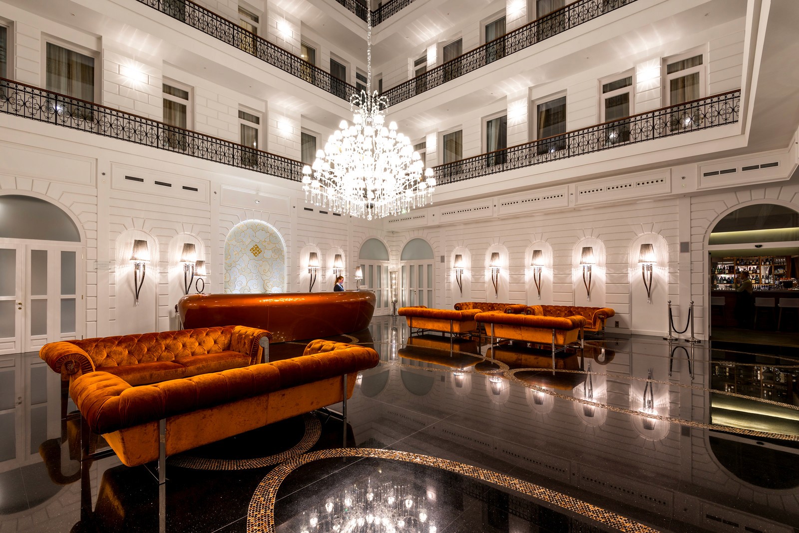 A grand hotel lobby with white walls, a large crystal chandelier, and plush orange velvet sofas in a wide hall.