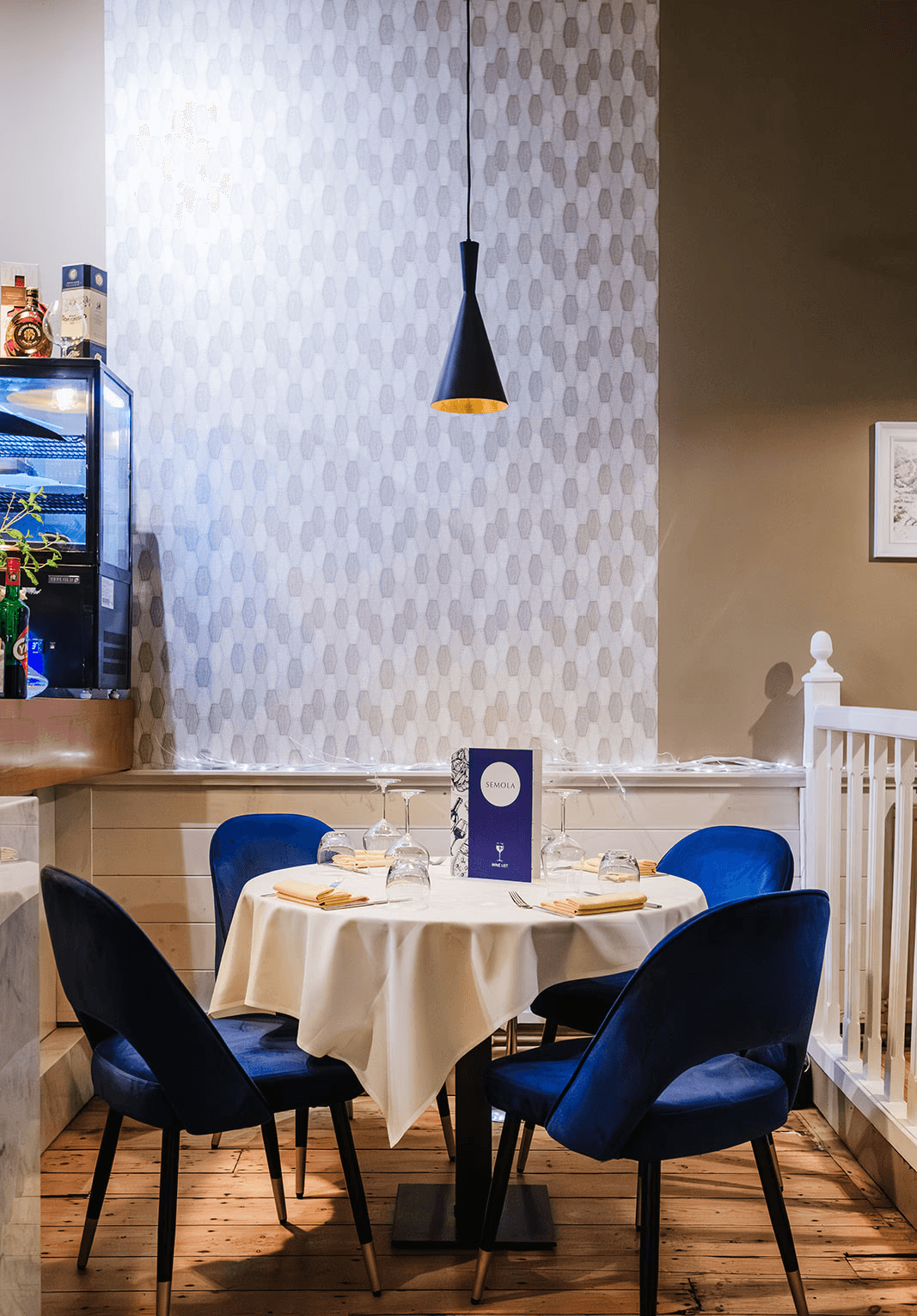 A neatly set round dining table with a cream tablecloth, surrounded by deep blue velvet chairs. A pendant light hangs above, and the table is arranged with folded napkins, upside-down glasses, cutlery and a Semola wine list placed at the centre, against a patterned grey wall with soft decorative lighting.