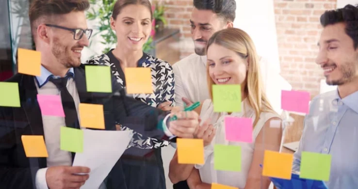Grupo de pessoas sorrindo diante de uma parede de vidro coberta de post-its coloridos, uma delas escrevendo