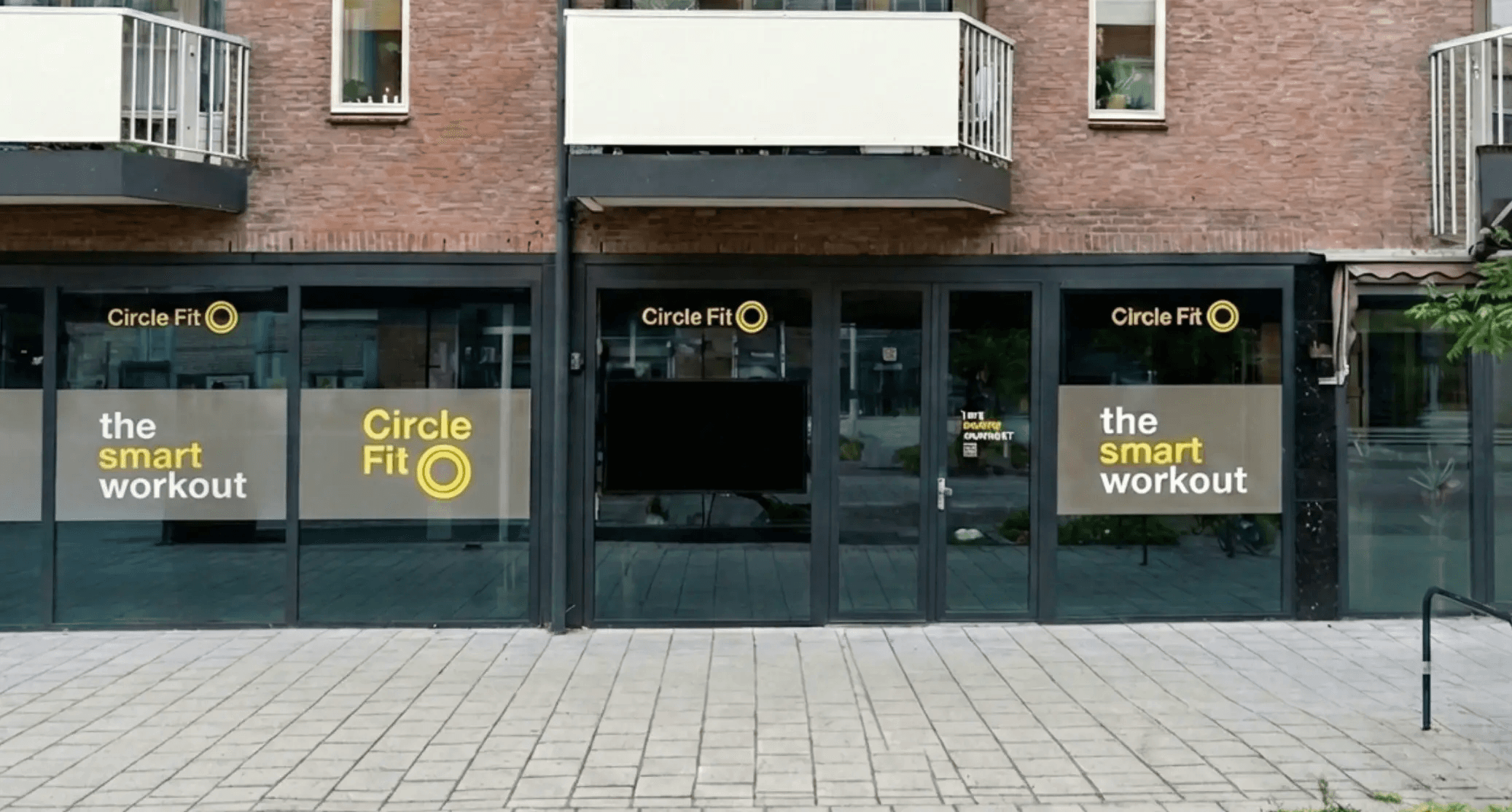 A Circle Fit hub with large windows displaying bright yellow signs and promotions, viewed from the sidewalk.