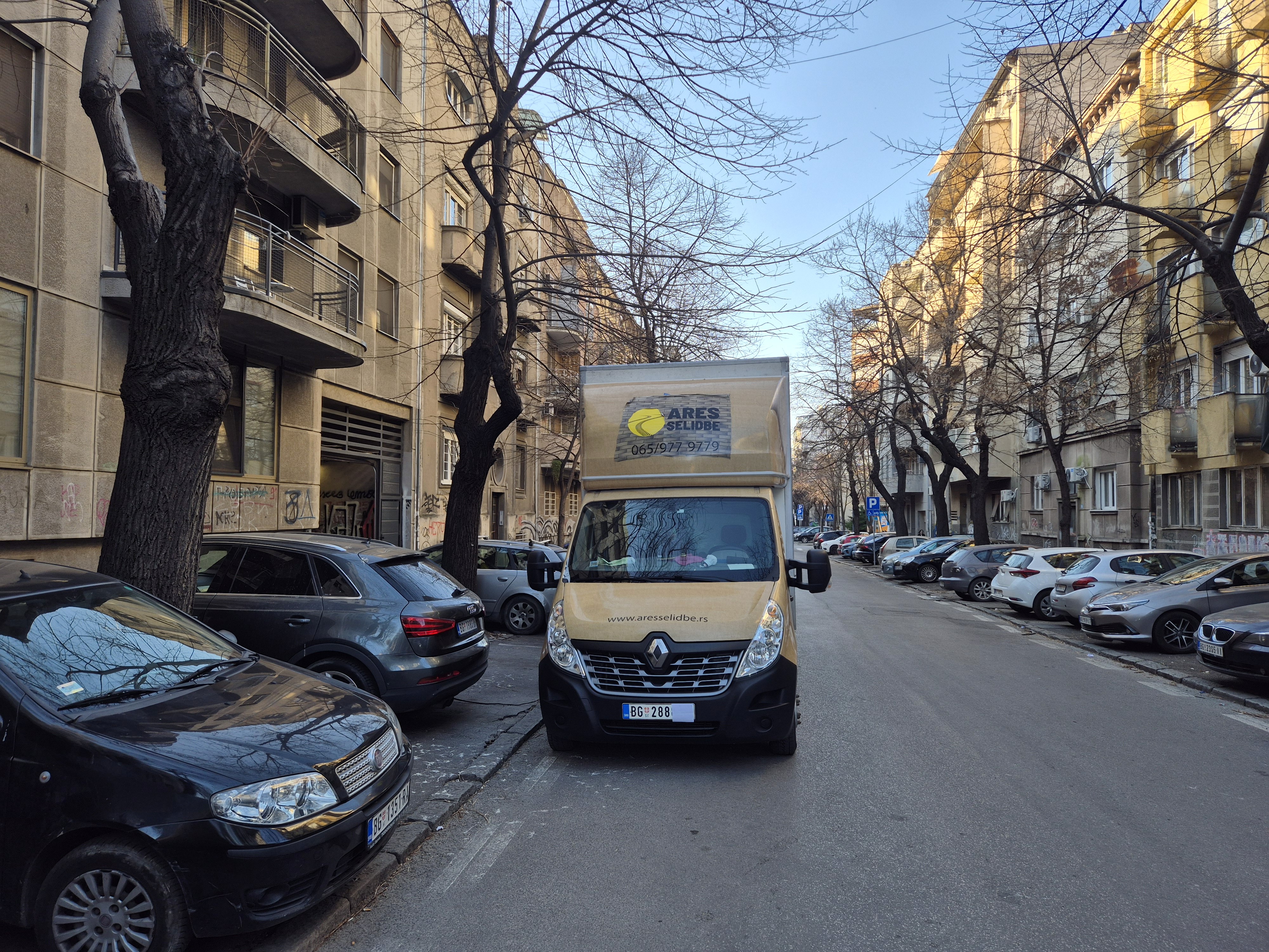 Kombi za selidbe parkiran u gradskoj ulici okruženoj zgradama i parkiranim automobilima.