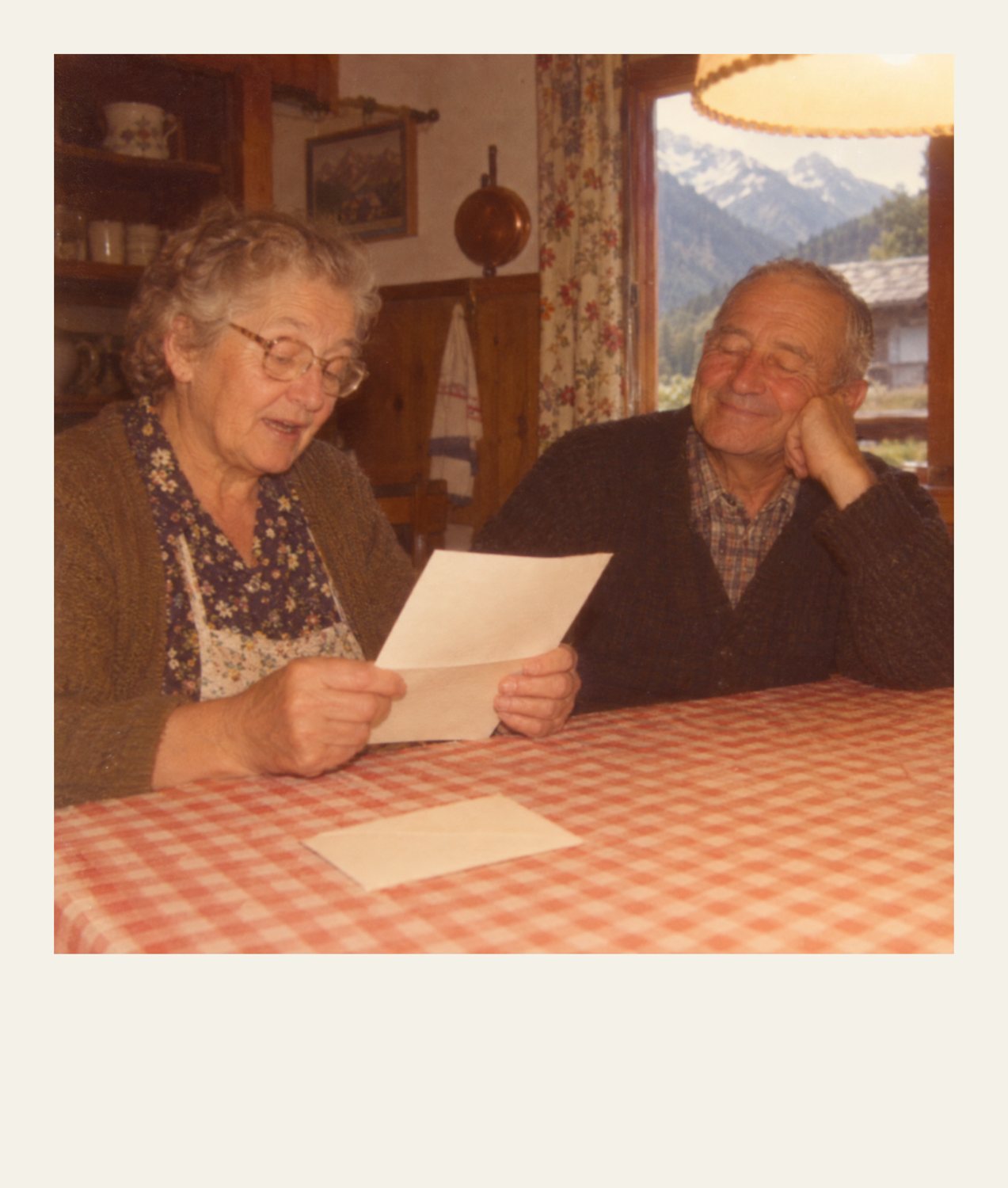 Polaroid of an elderly couple at a kitchen table; she reads aloud from a blank letter while he listens smiling