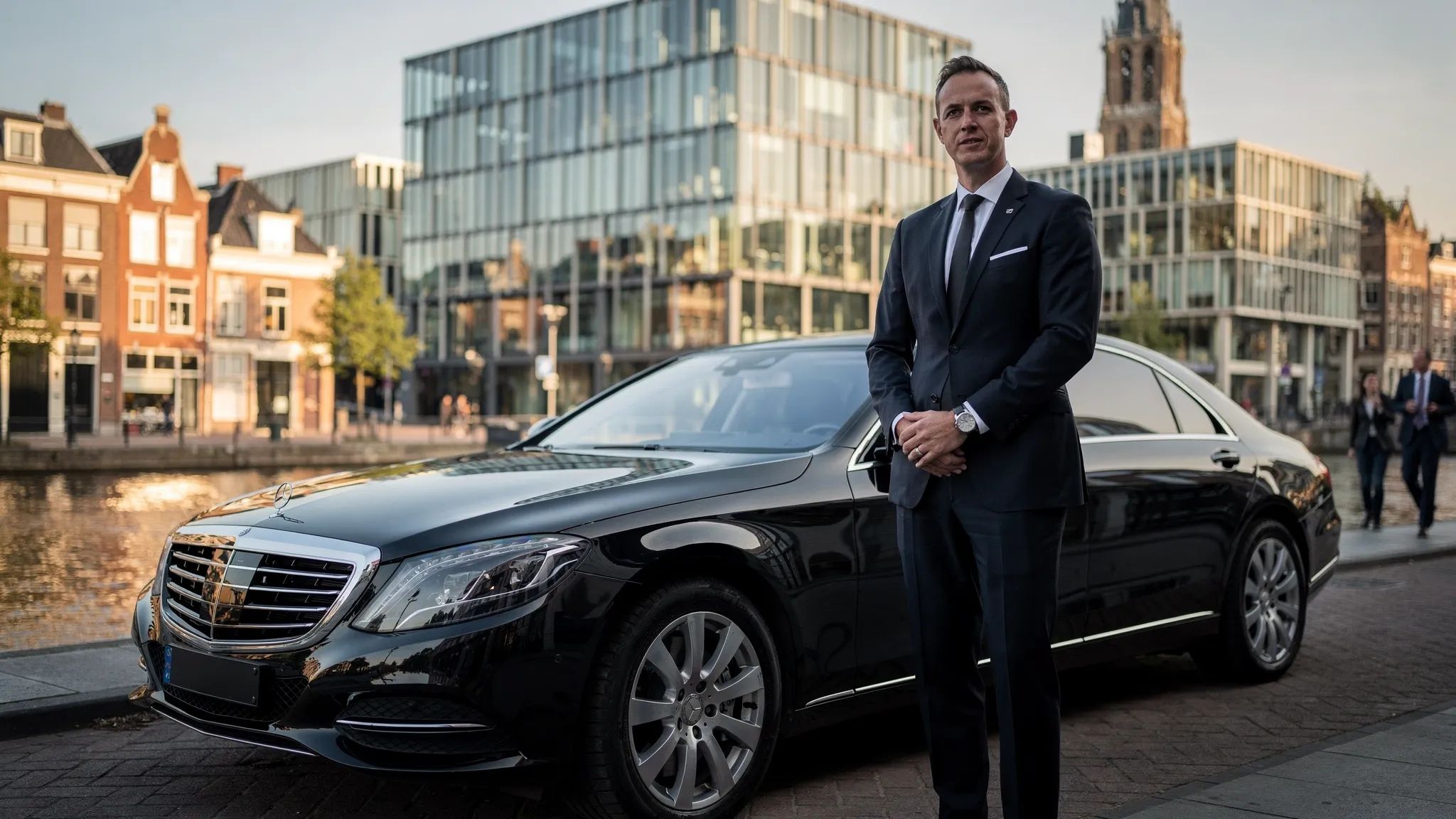 Een representatieve privéchauffeur wacht naast een luxe auto bij een zakelijke locatie in Utrecht, met op de achtergrond moderne kantoren en historische stadsdetails.
