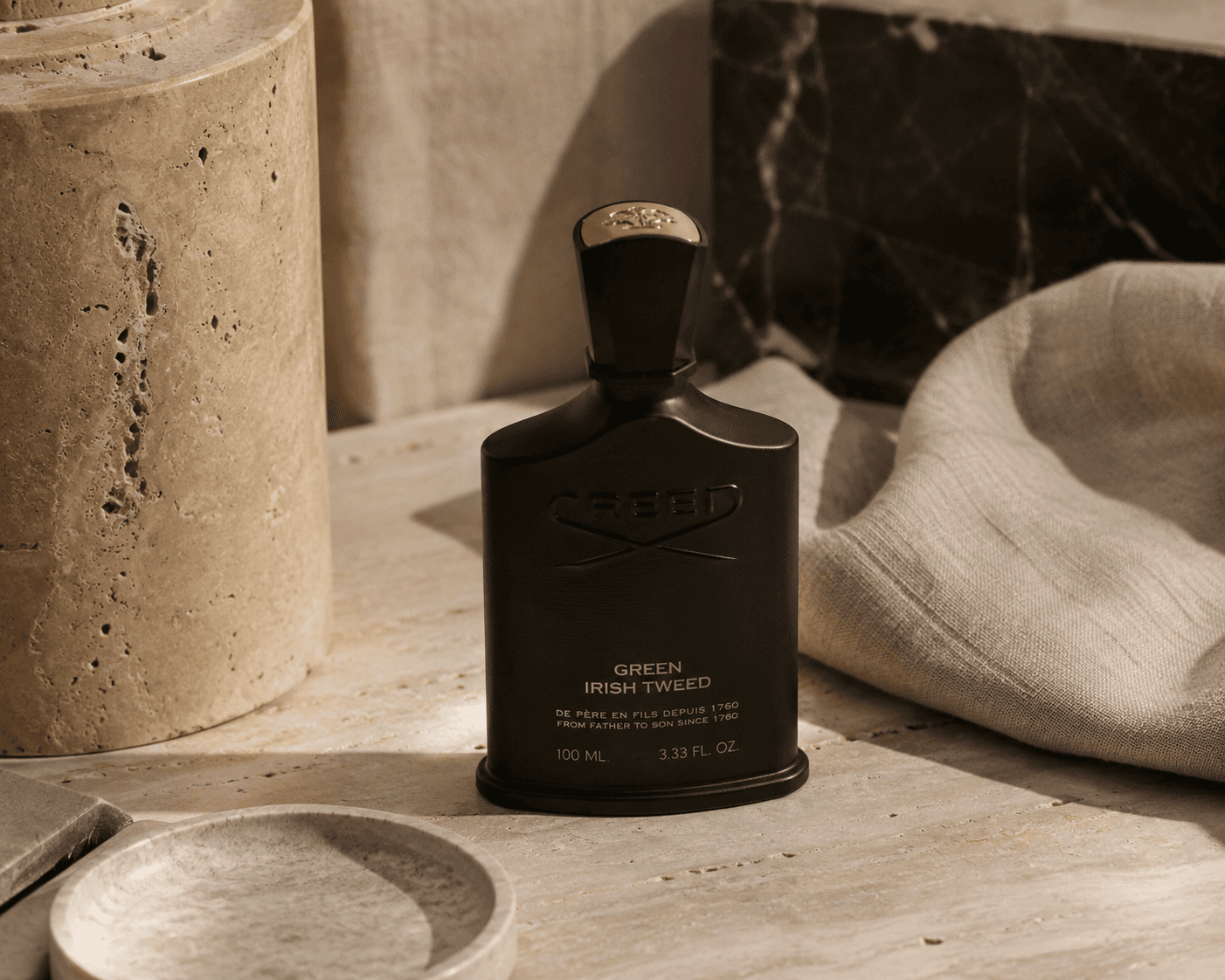 A matte black bottle of Creed Green Irish Tweed cologne standing upright on a beige travertine surface in direct warm sunlight. The bottle features embossed Creed lettering, gold typography, and a black cap topped with a gold crest. A textured stone cylinder, a shallow ceramic dish, dark marble, and draped linen cloth frame the scene.