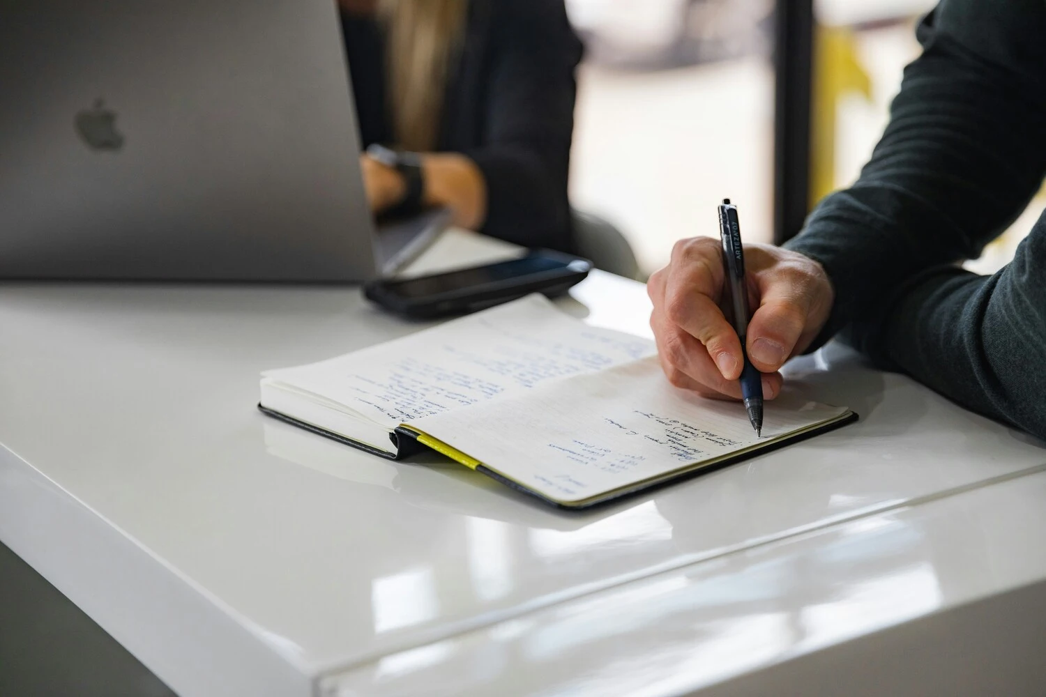 Close-up of a person's hand writing notes in a notebook at a desk.