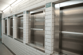Row of stainless steel dumbwaiter service lift doors set into white tiled wall in a commercial kitchen or hospital corridor — multiple units with landing call buttons visible