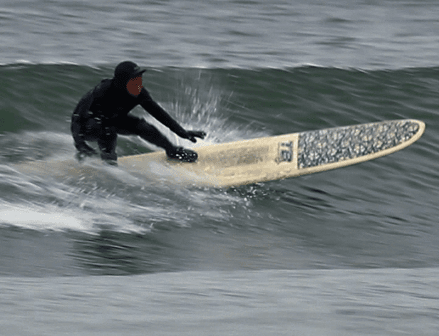 A person wearing a black wetsuit and hood is riding a cream-colored surfboard with a dark speckled pattern. The board is positioned diagonally on the water, with the front end pointing left and the back right. The surfer is crouched low on the board, with arms extended forward and knees bent. The water surface is dark and slightly choppy, with small waves visible around the board. The image is slightly blurred, likely due to motion.