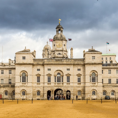 Historic multi-arched stone building with a clock tower and flags, set against a cloudy sky, with an open courtyard in front.