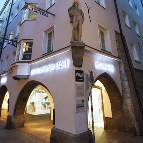 Exterior da loja Swarovski iluminada com portas em arco, grandes janelas e uma estátua no canto do edifício ao anoitecer.