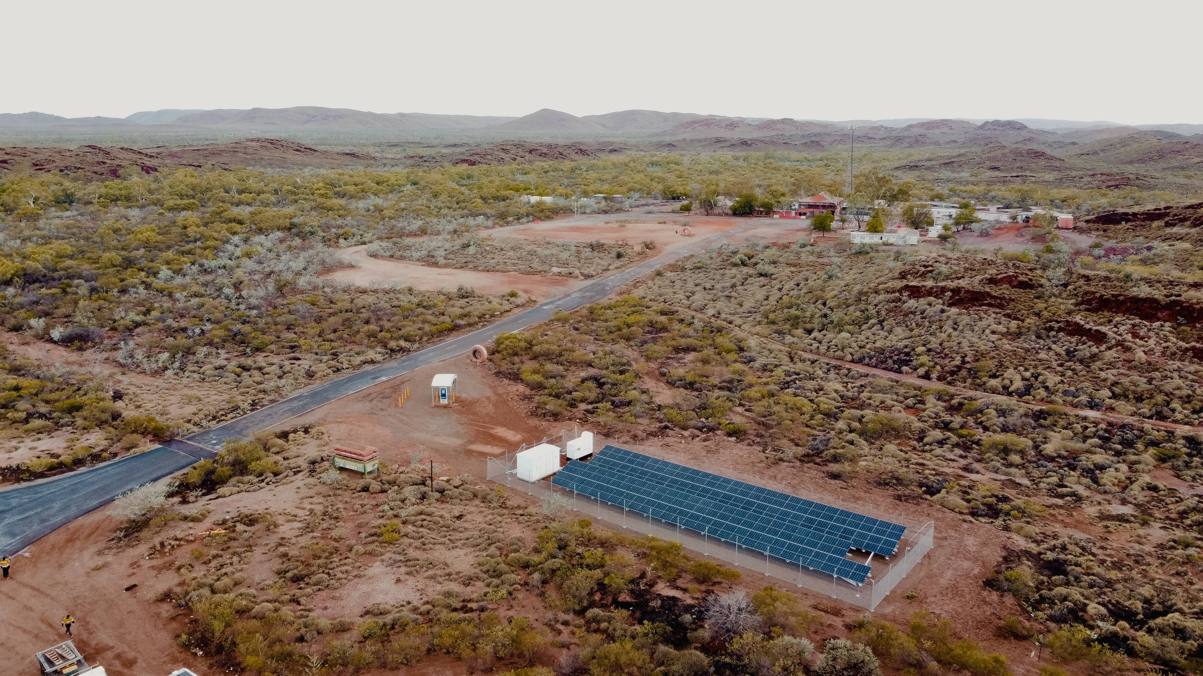 Drone aerial view of a large solar panel array and energy storage containers in a remote, arid Australian landscape with hills and sparse vegetation in the background.