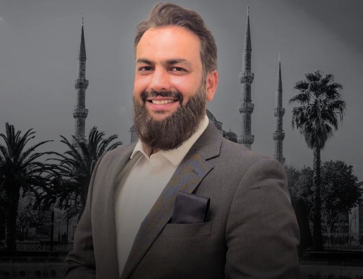 A young man with light brown hair is wearing a black suit jacket and white shirt, standing in a softly lit office setting, conveying professionalism.