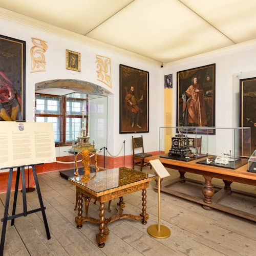 A museum room with framed portraits, display cases containing intricate models, an ornate table, and informational boards.