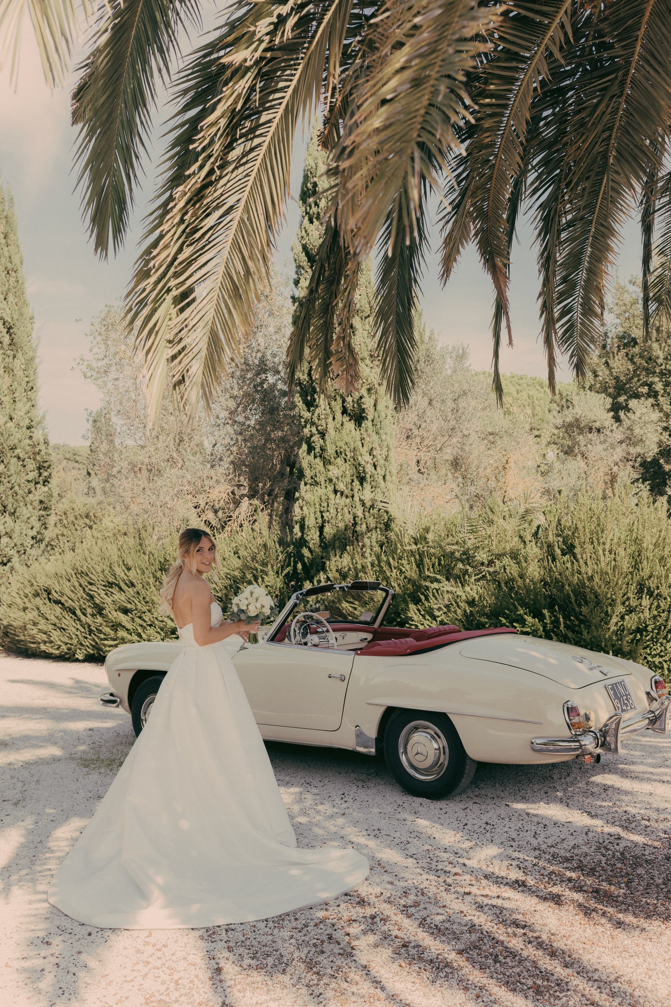 Sposa in abito bianco accanto a Mercedes d'epoca decappottabile a Bolgheri, tra palmizi e luce dorata