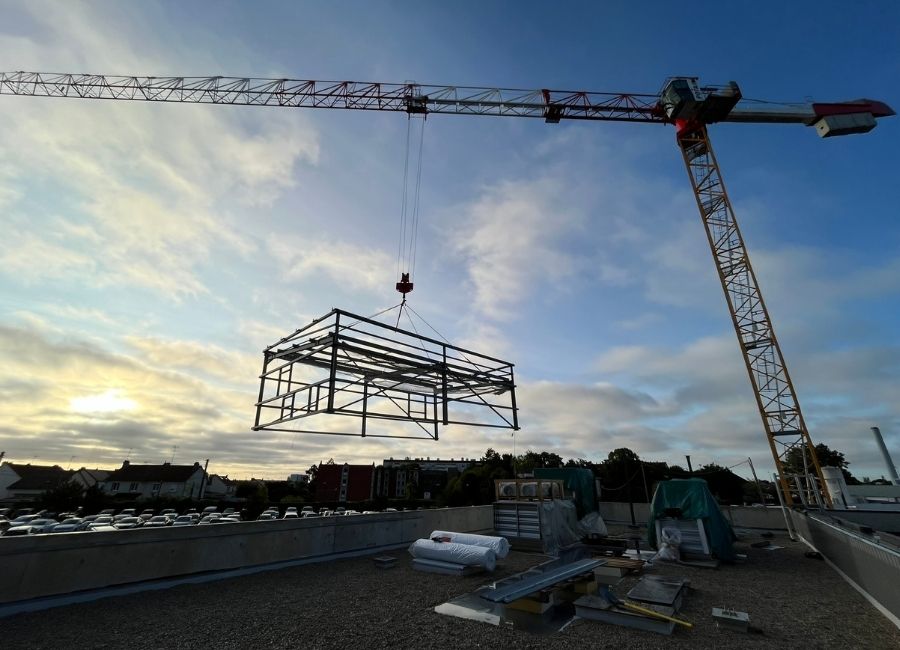 Structure métallique en cours de levage par grue sur un chantier urbain au coucher du soleil.
