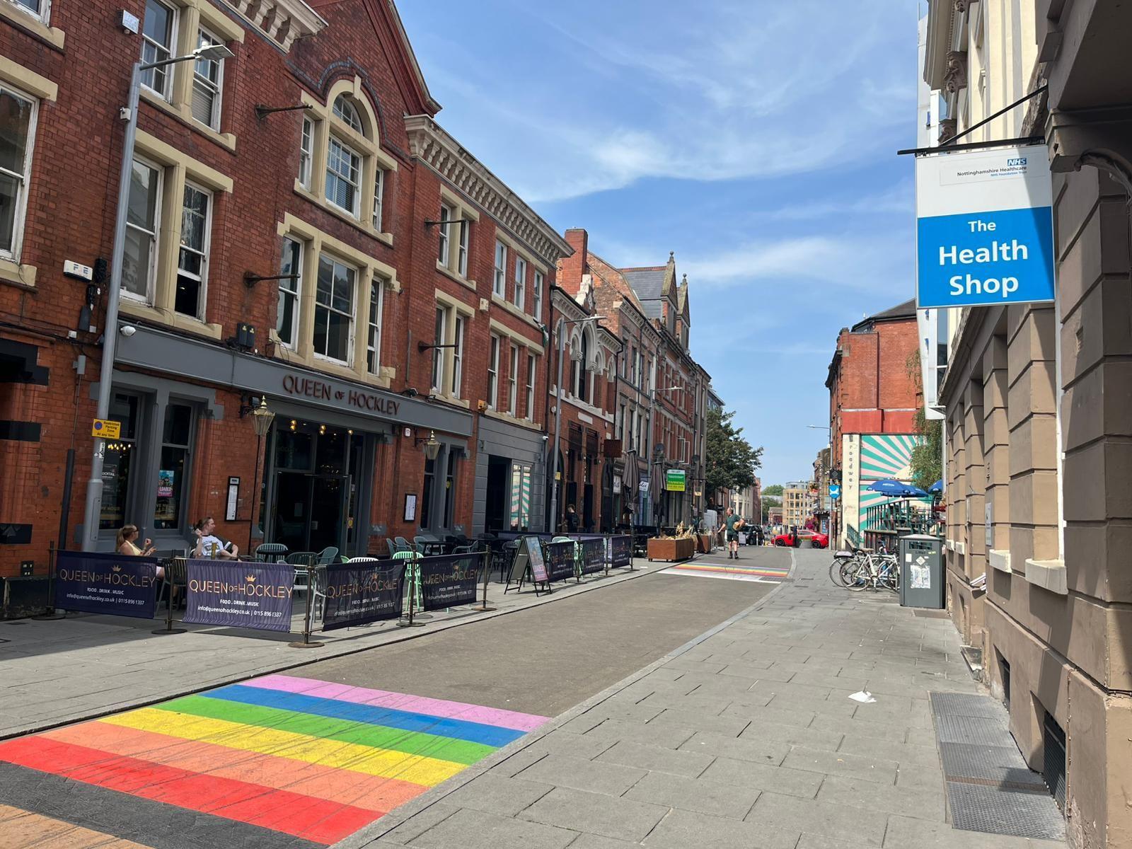 A street in Nottingham on a sunny day. In the foreground there is a rainbow crossing on the floor. On the right hand side there is a blue sign which says 'health shop'. In the background are red brick buildings 