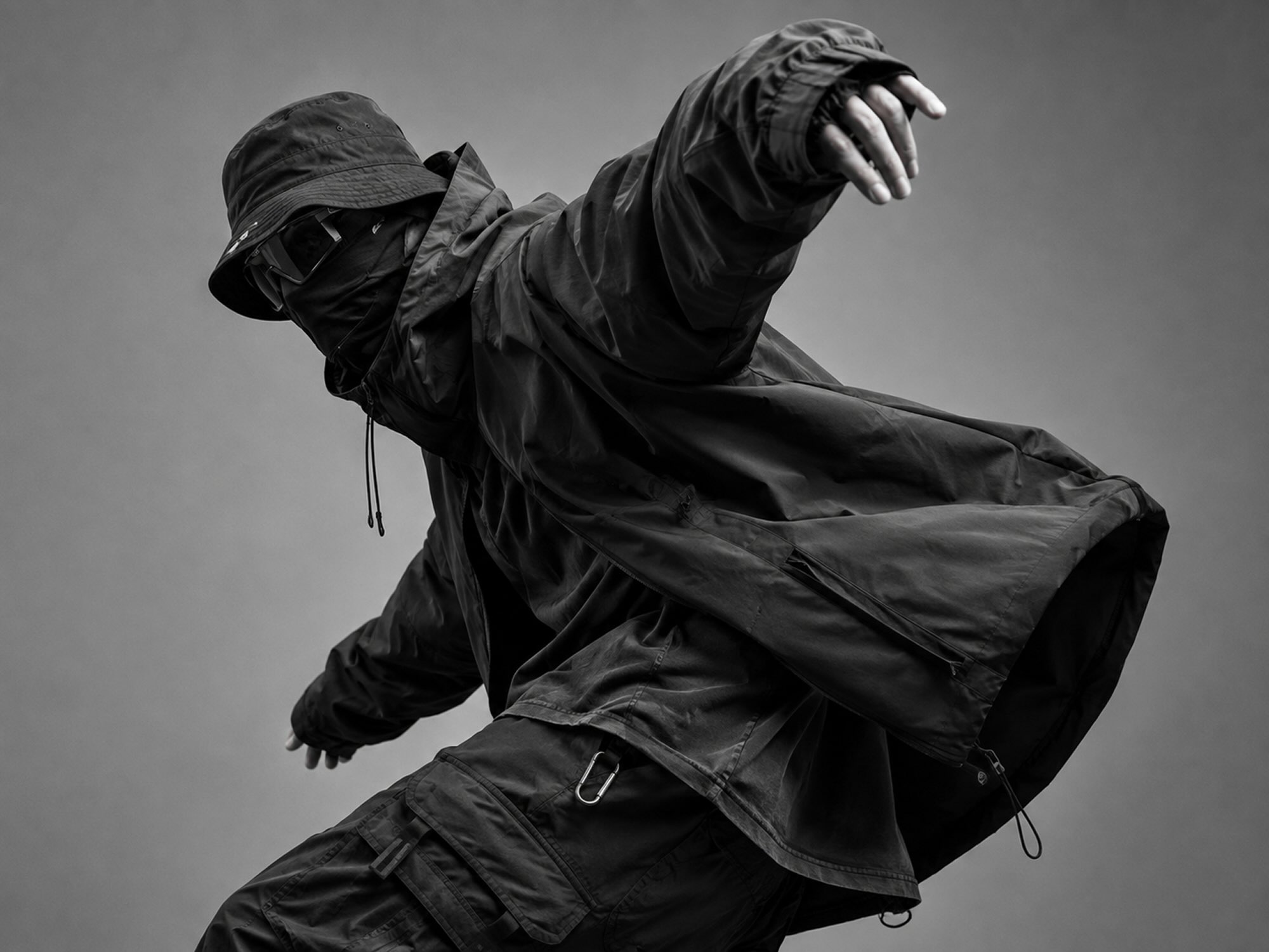 Black-and-white image of a person in dark clothing, wearing a cap and face covering, leaning forward with one arm extended.