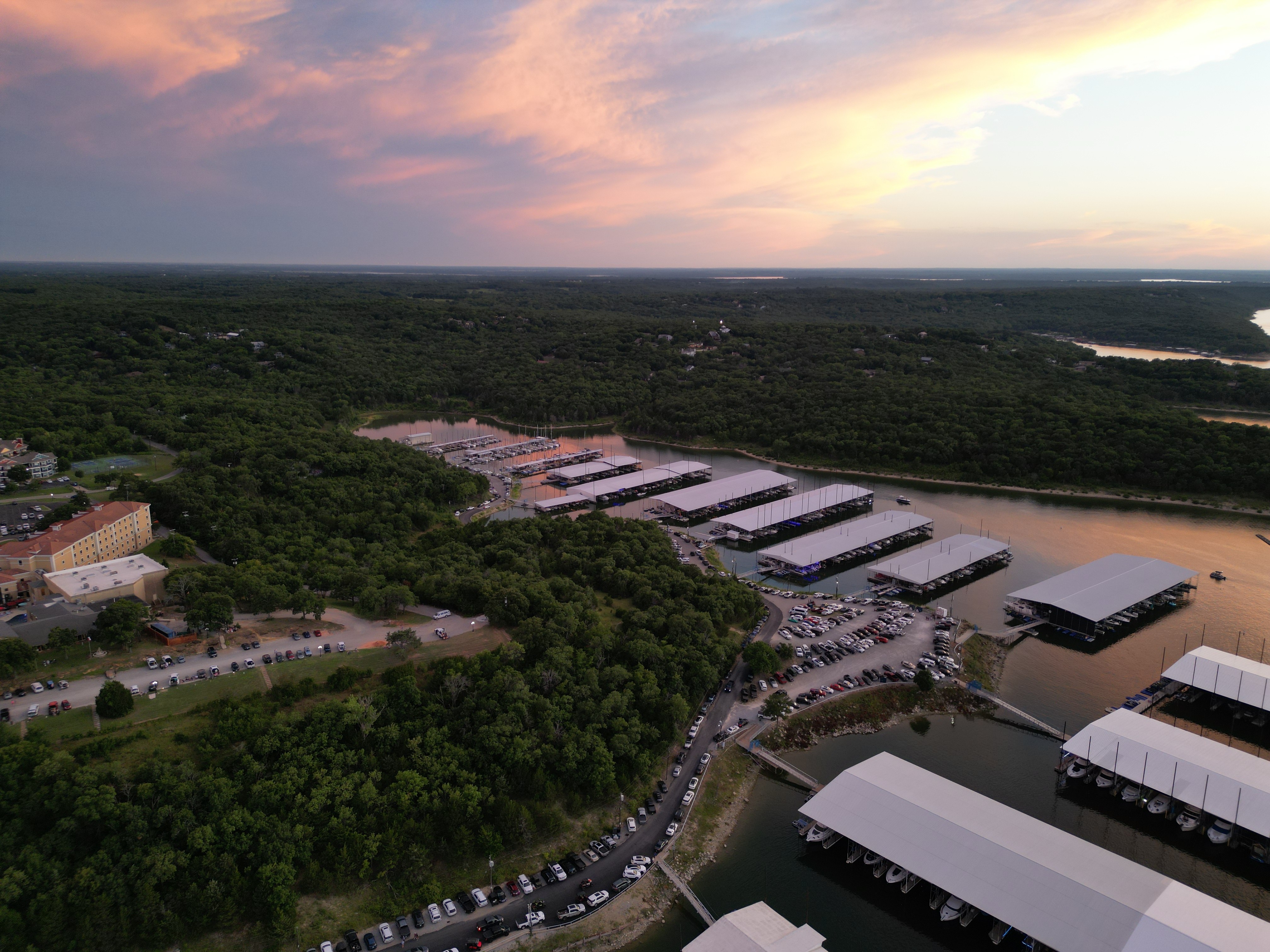 Aerial view of a marina with multiple covered boat docks surrounded by lush greenery, set along a winding body of water under a clear blue sky.