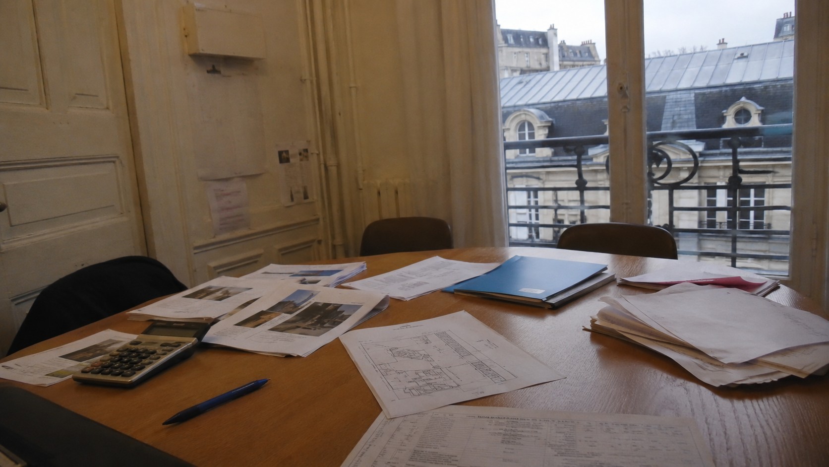 Vote de travaux de toiture en assemblée générale de copropriété à Paris avec devis, plans techniques, calculatrice et documents du syndic sur une table.