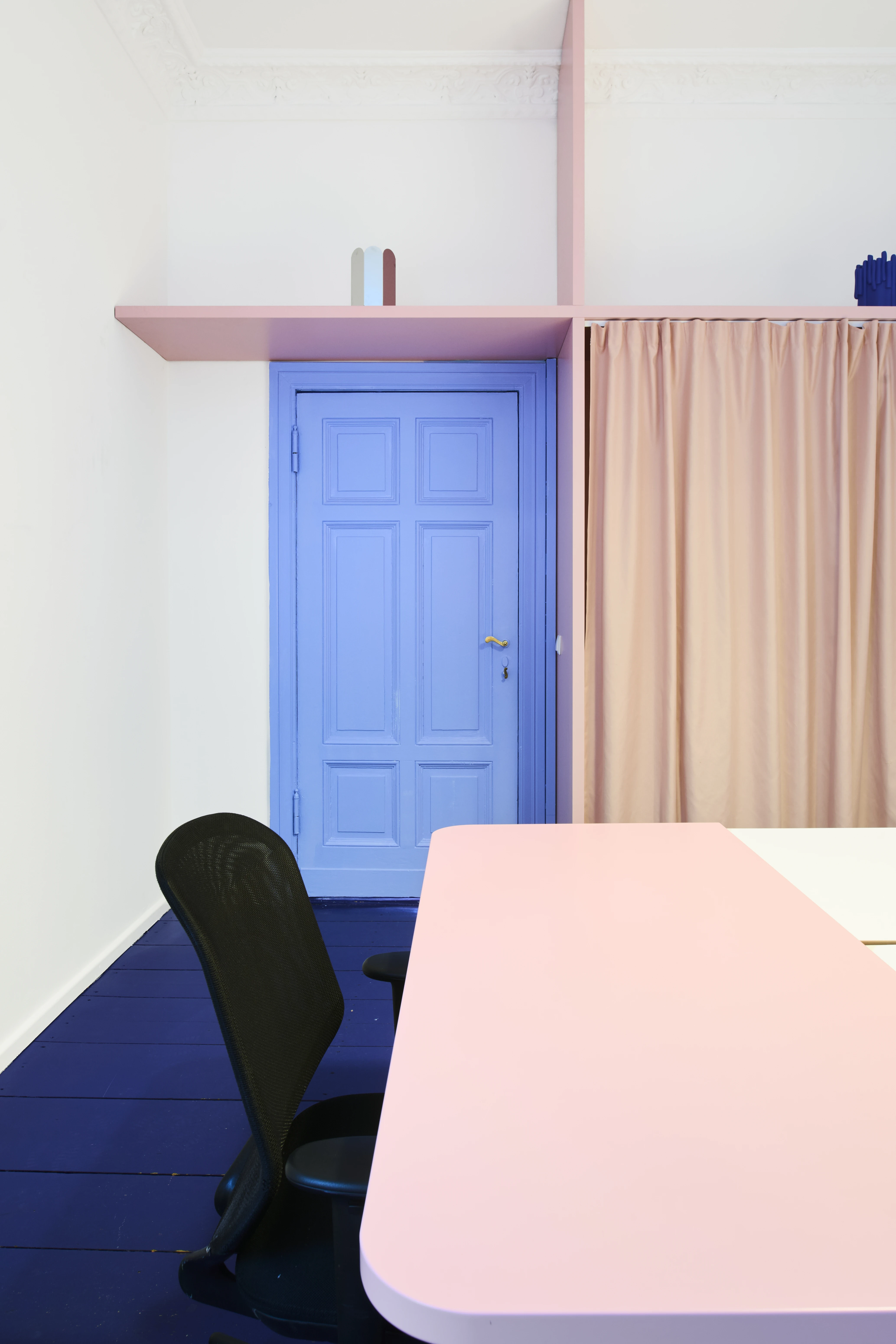 Conference room corner with blue panelled door, pink shelf, beige curtain and rose table.