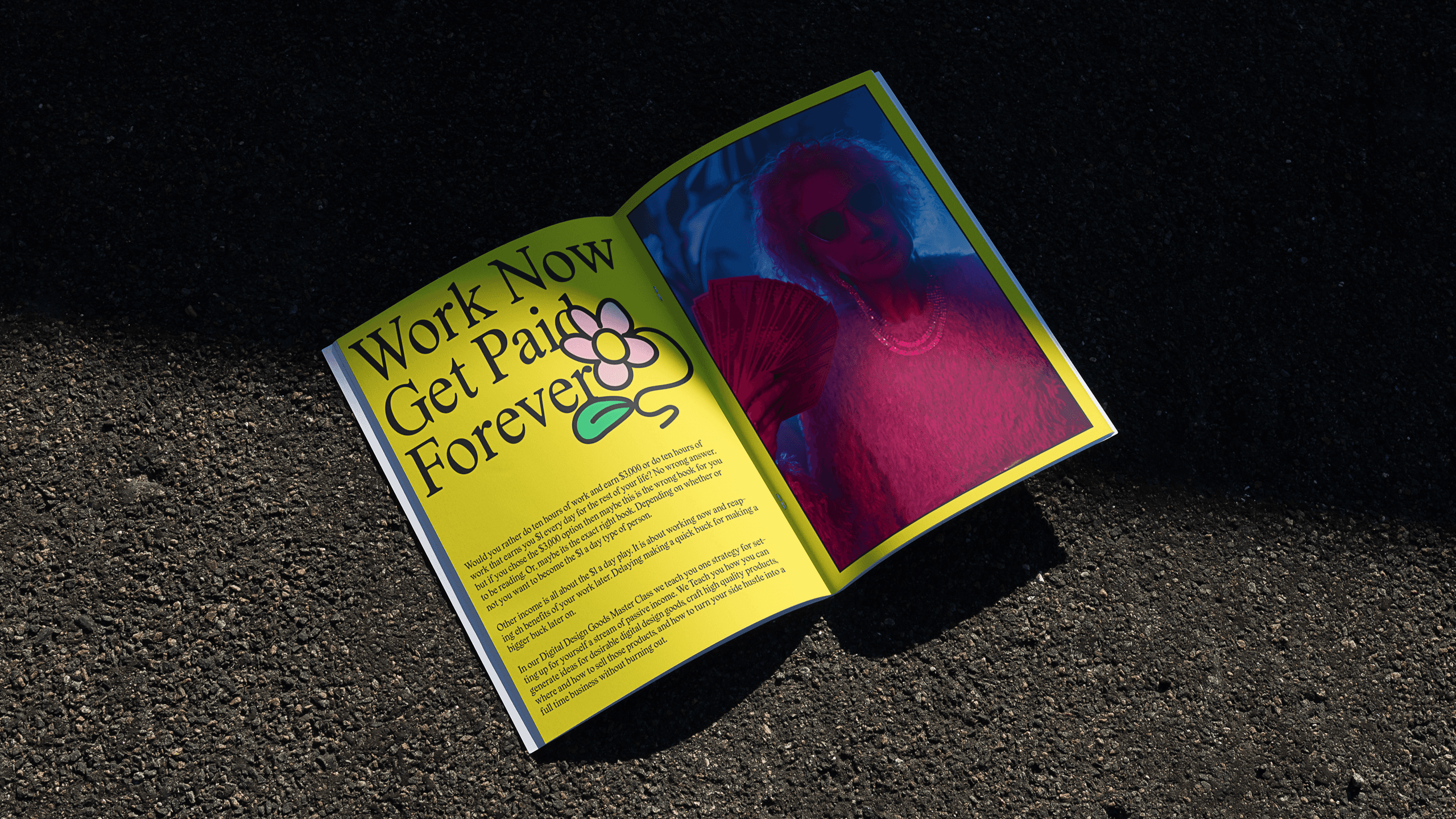The image shows an open book lying on a dark asphalt surface. The left page is yellow and displays the text "Work Now Get Paid Forever" with a small flower illustration near the word "Forever". The right page is blue and features a photograph of a person wearing sunglasses and a pink top. The book is positioned diagonally, with the left page partially covering the right page. The asphalt surface has small aggregates visible, and the lighting creates shadows, particularly to the right of the book.