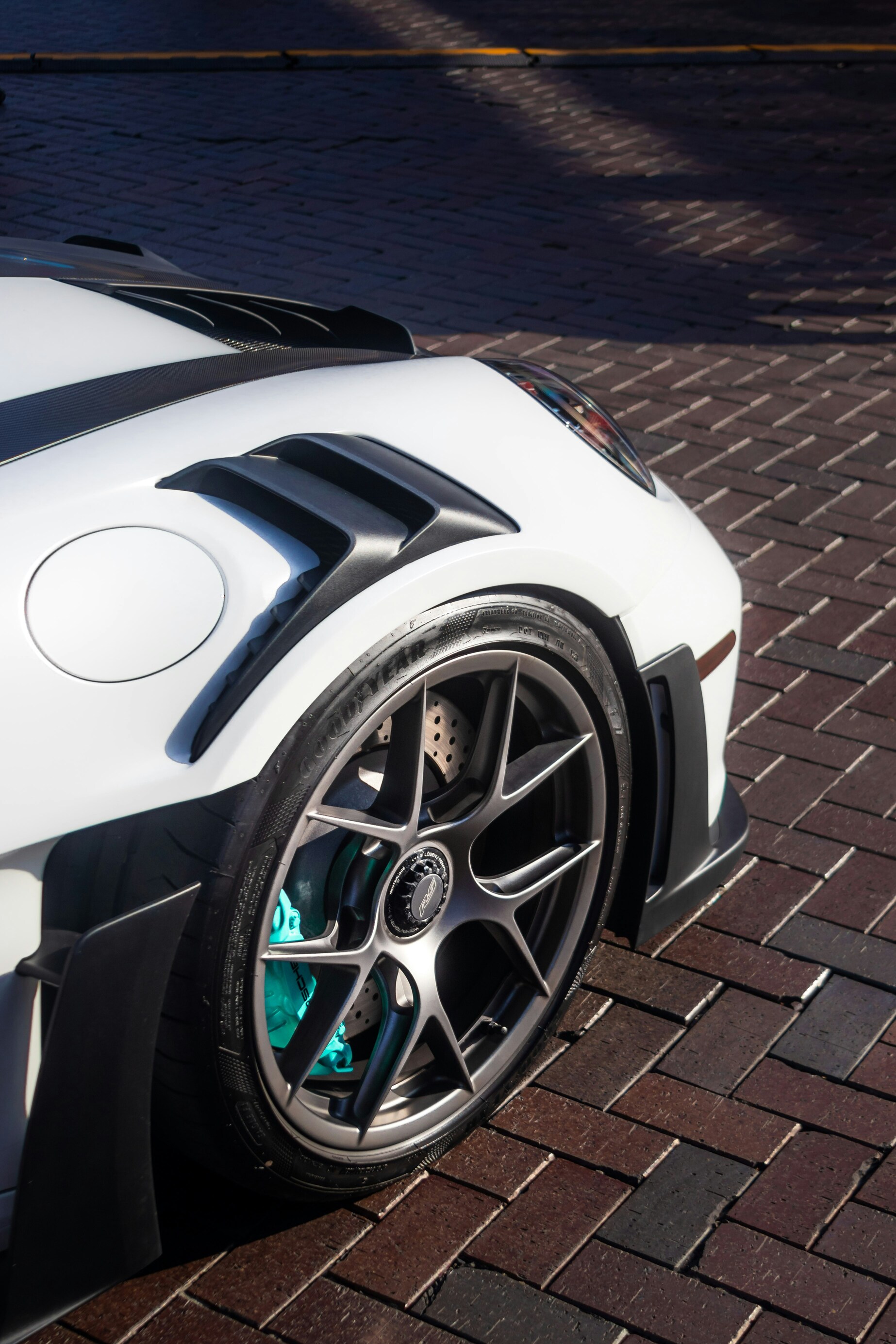 The image showcases the close-up view of a sleek, white sports car with detailed black wheel arches and a stylish alloy wheel adorned with teal-colored brake calipers, parked on a sunlit brick street.