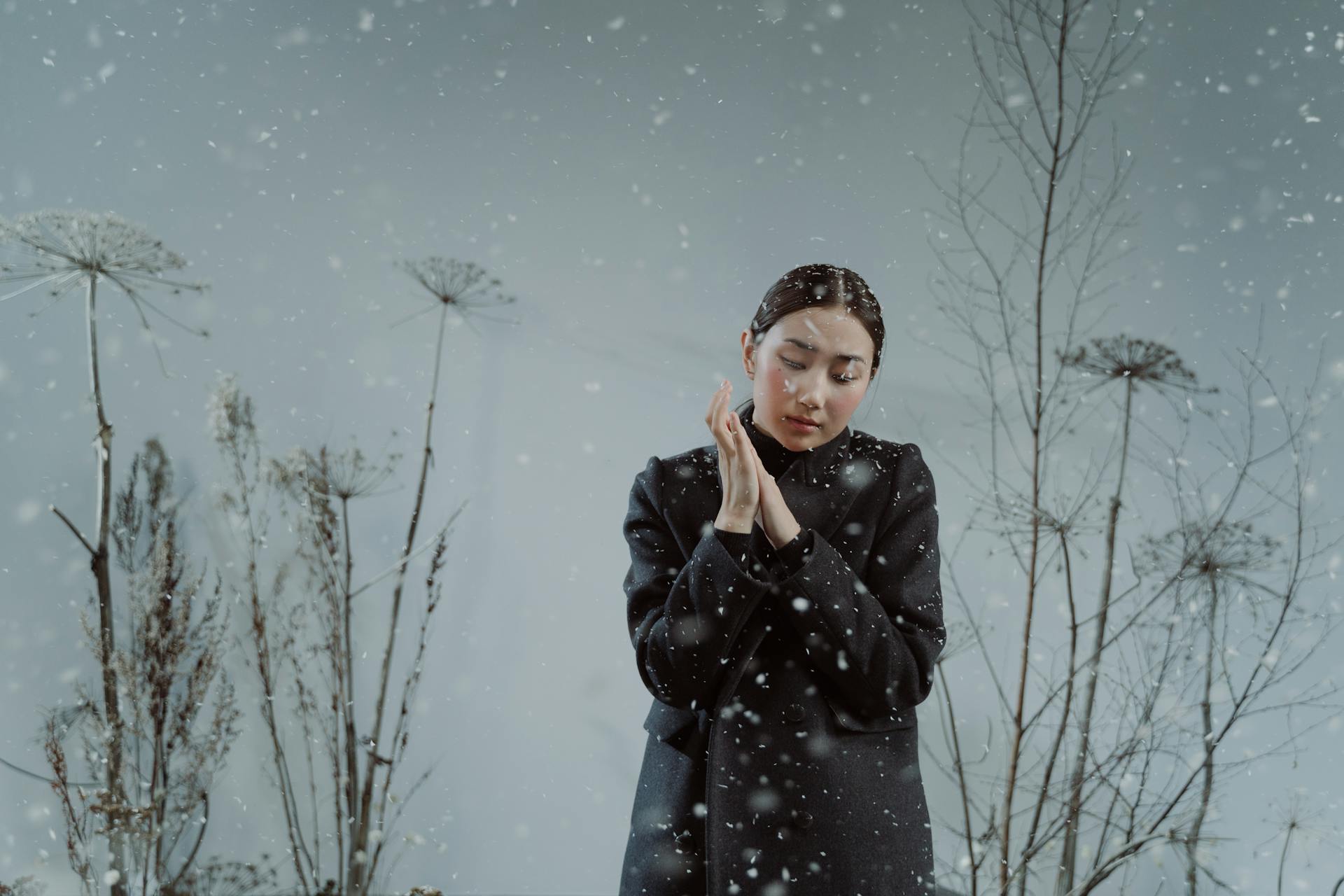 A woman in a dark grey coat stands with her eyes closed and hands pressed together in prayer as snow falls around her amidst dried, tall plants.