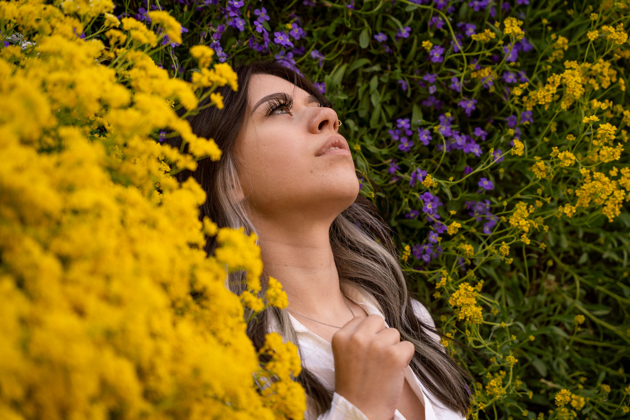 A woman sitting with flowers, all around her, gazing up into the sky