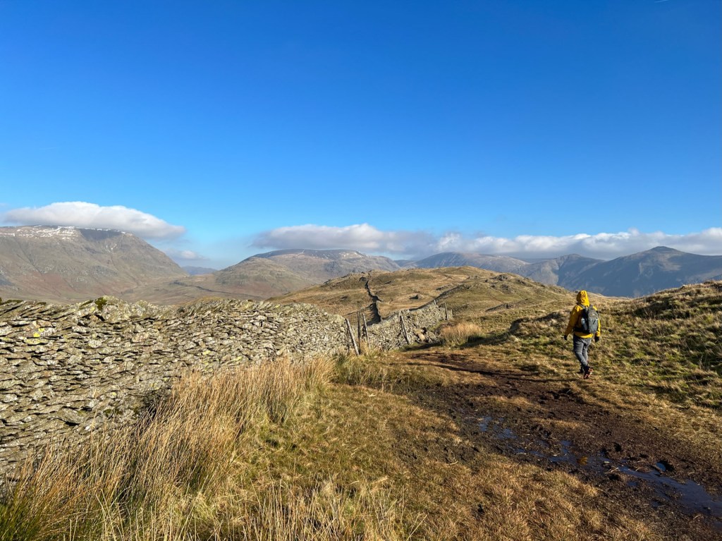 Martin walking around a boggy marsh on the way to Wansfell. A stone wall trails off into the distance on the left, and the surround fells can be seen in the background. Clear blue skies with just a few clouds sitting on top of the distant fells.