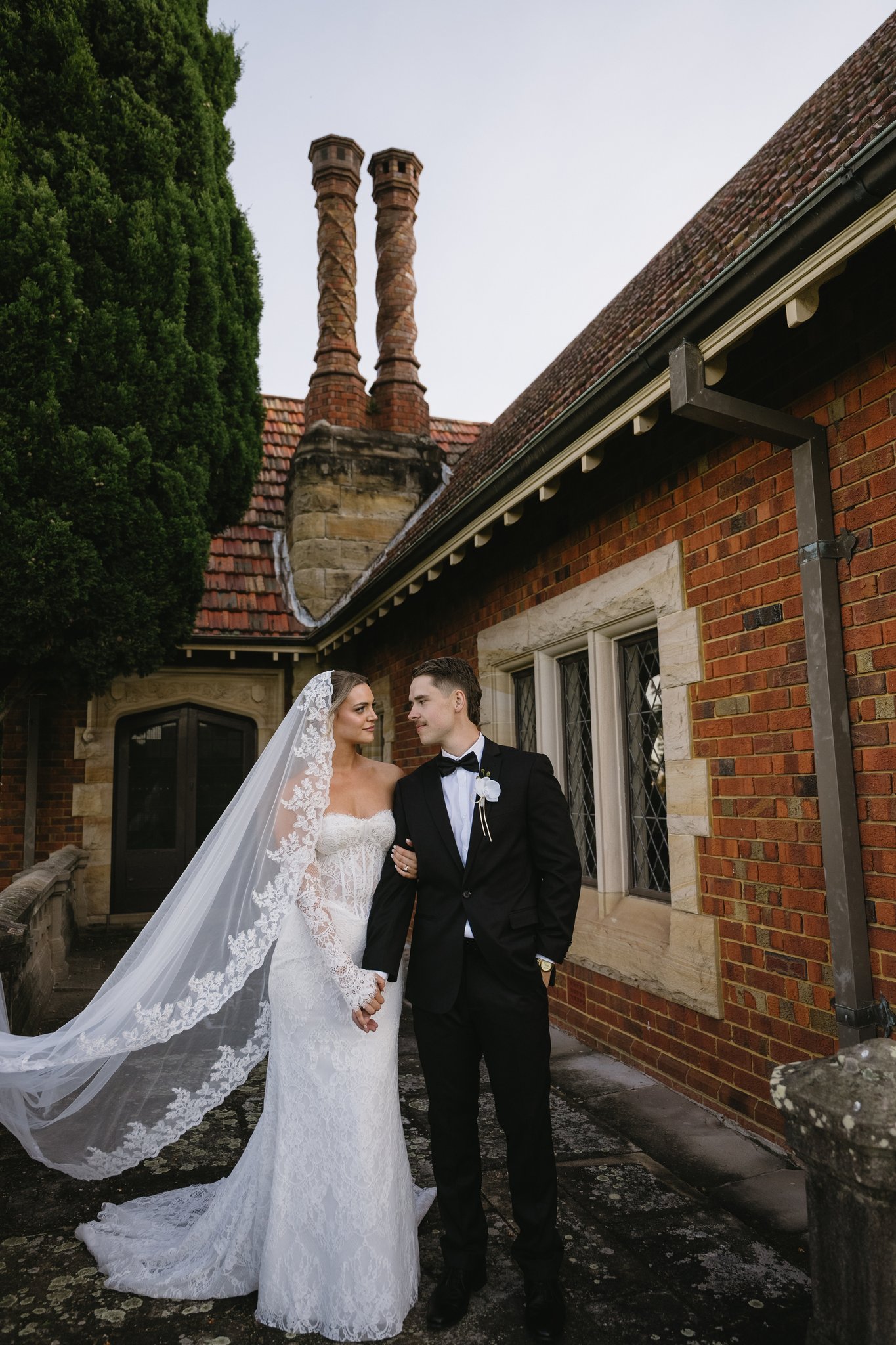 Bride in lace gown and veil with groom in tuxedo standing beside a brick building with tall twisted chimneys.