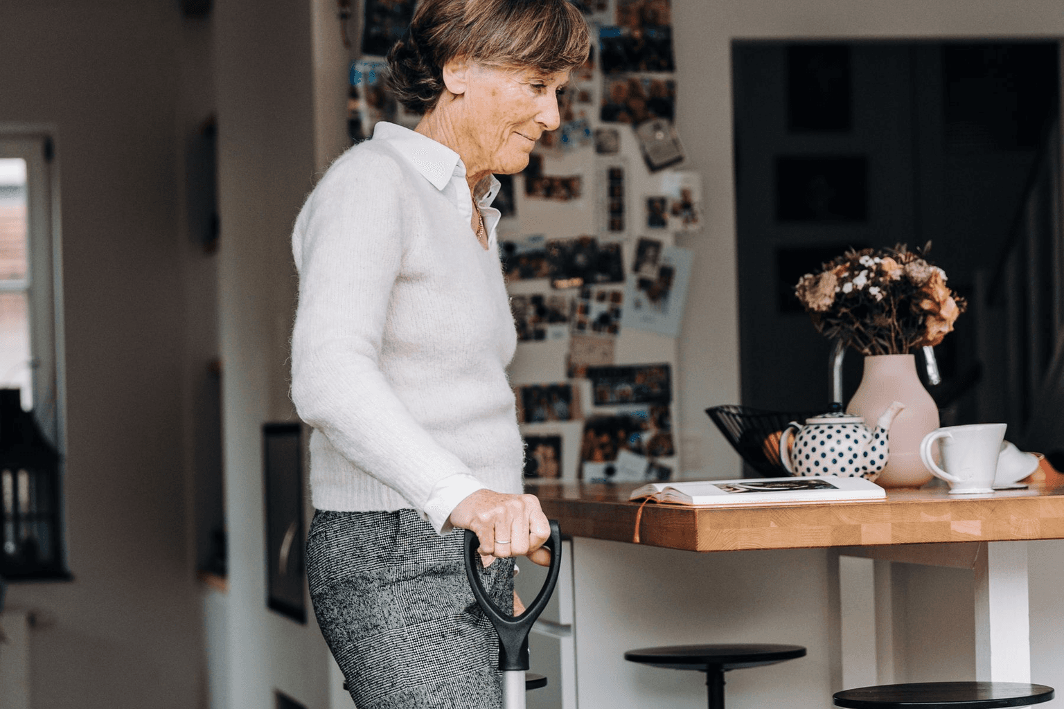 An older woman moving through her own kitchen with a walking frame, her tea and flowers on the counter.