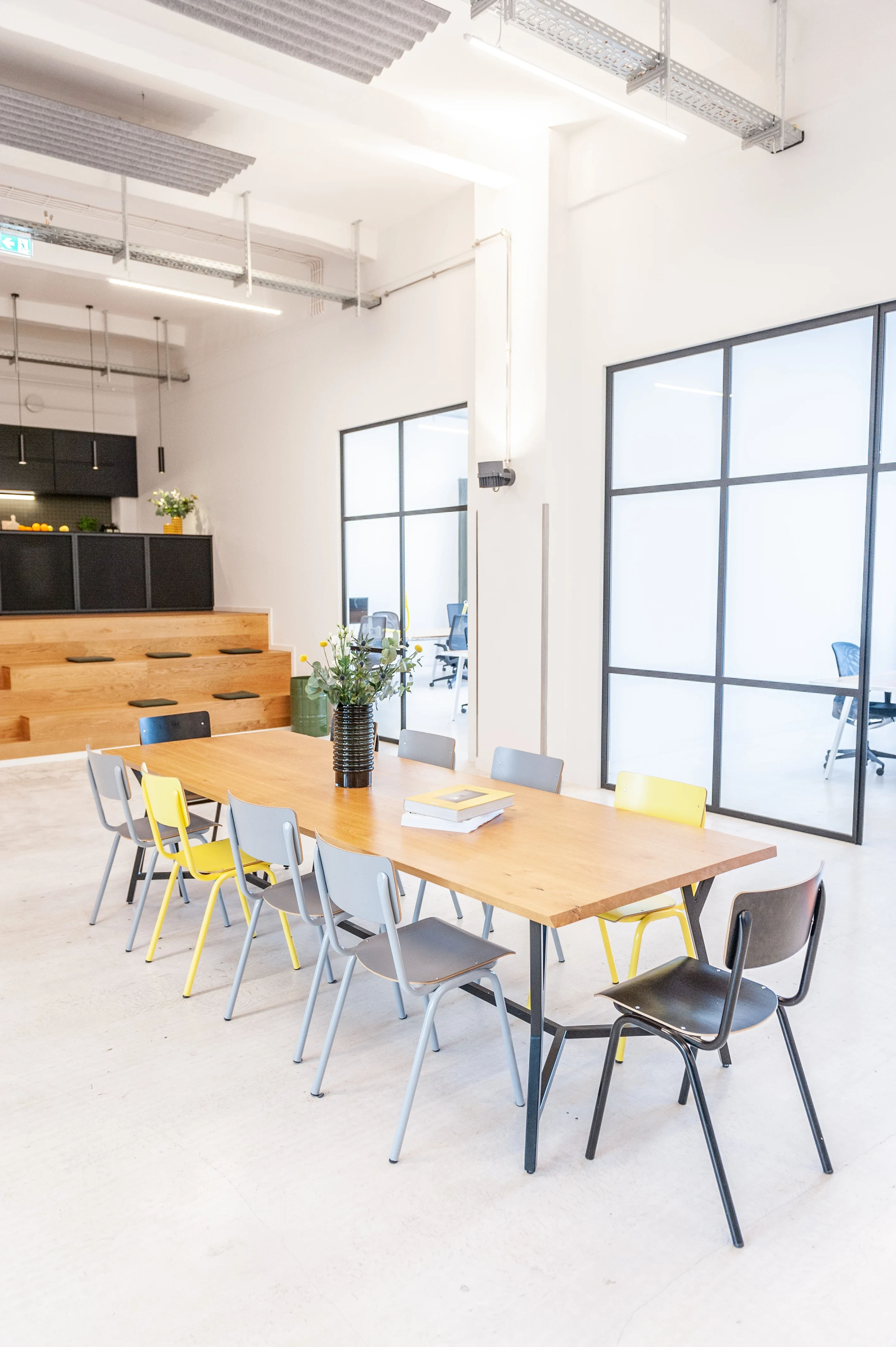 Open meeting area at LA RED with a long wooden table surrounded by grey and yellow school-style chairs, a tiered wooden seating staircase and dark kitchen counter in the background.