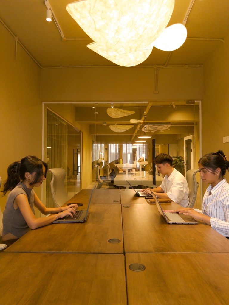 A modern coworking space featuring three people focused on their laptops at a large wooden table. The room is adorned with warm earthy tones and a unique lighting fixture, creating a comfortable yet productive atmosphere.