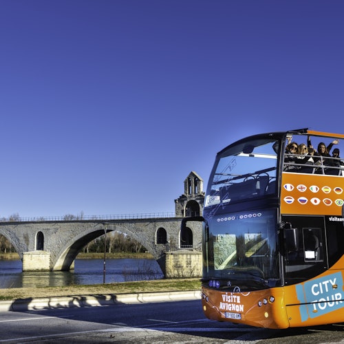Dvoupodlažní turistický autobus s nápisem "Visite Avignon" vedle historického kamenného mostu přes řeku za jasného dne.