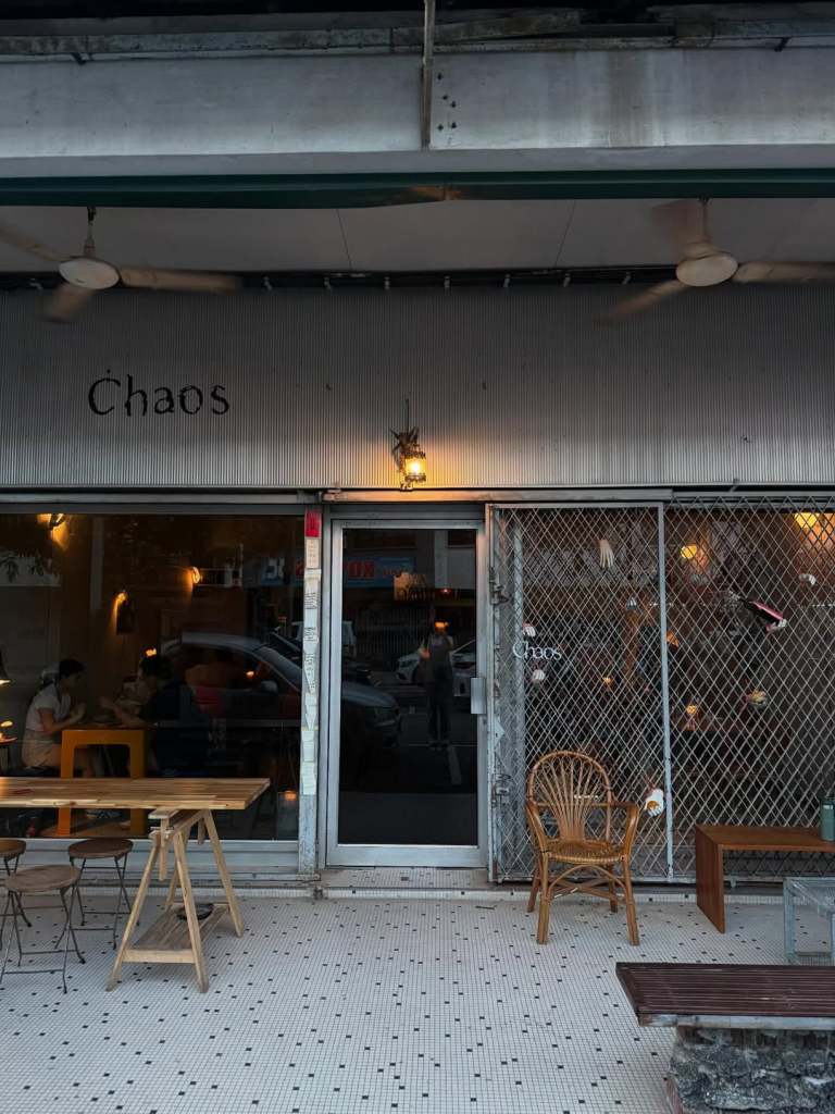 Front view of a café named 'Chaos' featuring a glass door, outdoor seating with wooden tables, and a decorative façade with warm lighting.