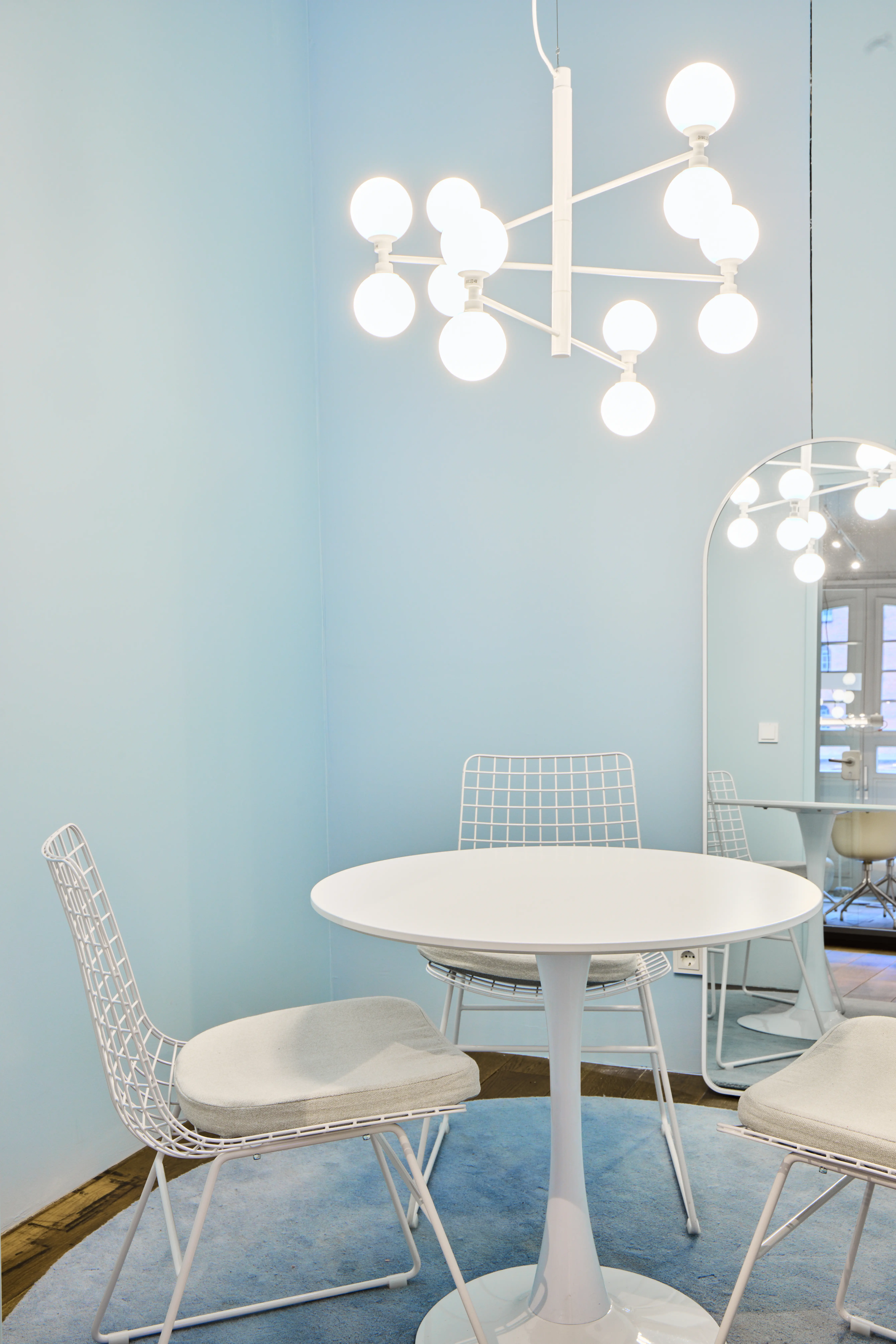 Light blue meeting corner at INTERMATE OFFICE HAMBURG with a round white tulip table, white wire chairs with grey cushions, a white sphere chandelier, an arched floor mirror, and a round light blue rug.