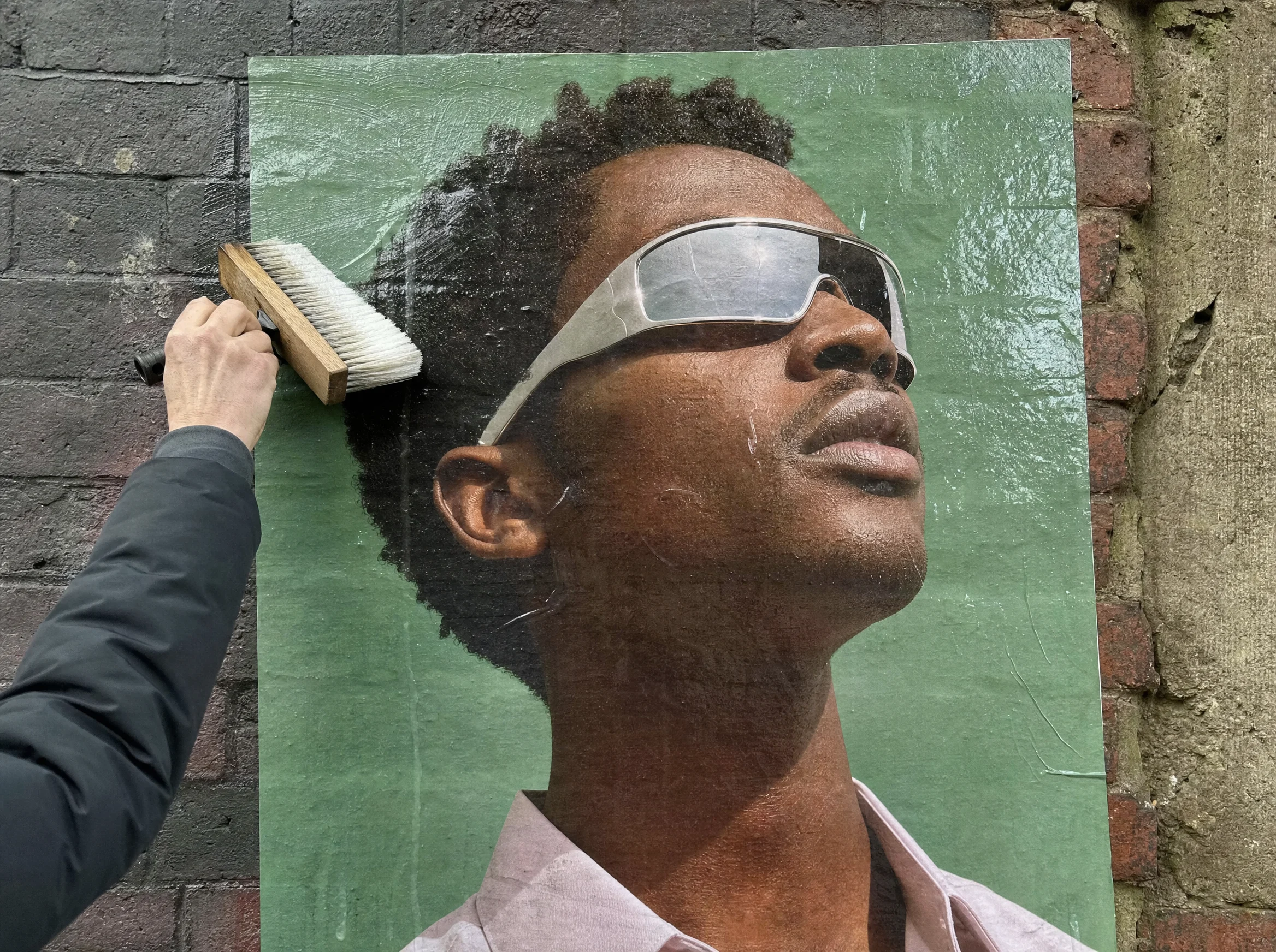 Hand pasting a Ferro poster of a man in chrome sunglasses onto a brick wall