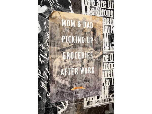 The image shows a weathered poster affixed to a dark wall. The poster features white text reading "MOM & DAD PICKING UP GROCERIES AFTER WORK" in a distressed font. The background of the poster has a faded, vintage appearance. A small, orange-toned sticker or label is visible near the bottom center of the poster. The wall behind the poster is dark and textured, with portions of other posters or signs visible to the right, though the text on them is not legible.