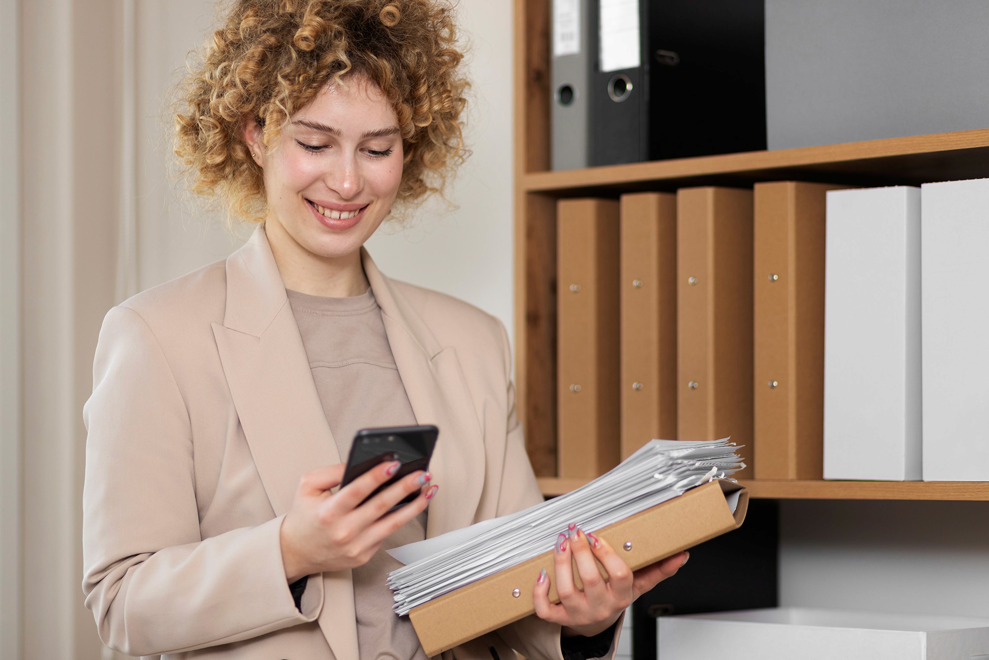 Smiling woman holding documents and using her phone in an office