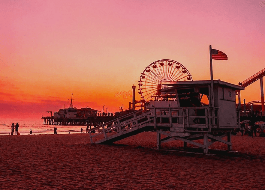 A dreamy pink and orange sunset over a sandy beach with a lifeguard  tower and Ferris wheel silhouetted on the pier — the warm, film-inspired  light at the heart of Retrica 9.2's Prism and Makeup features