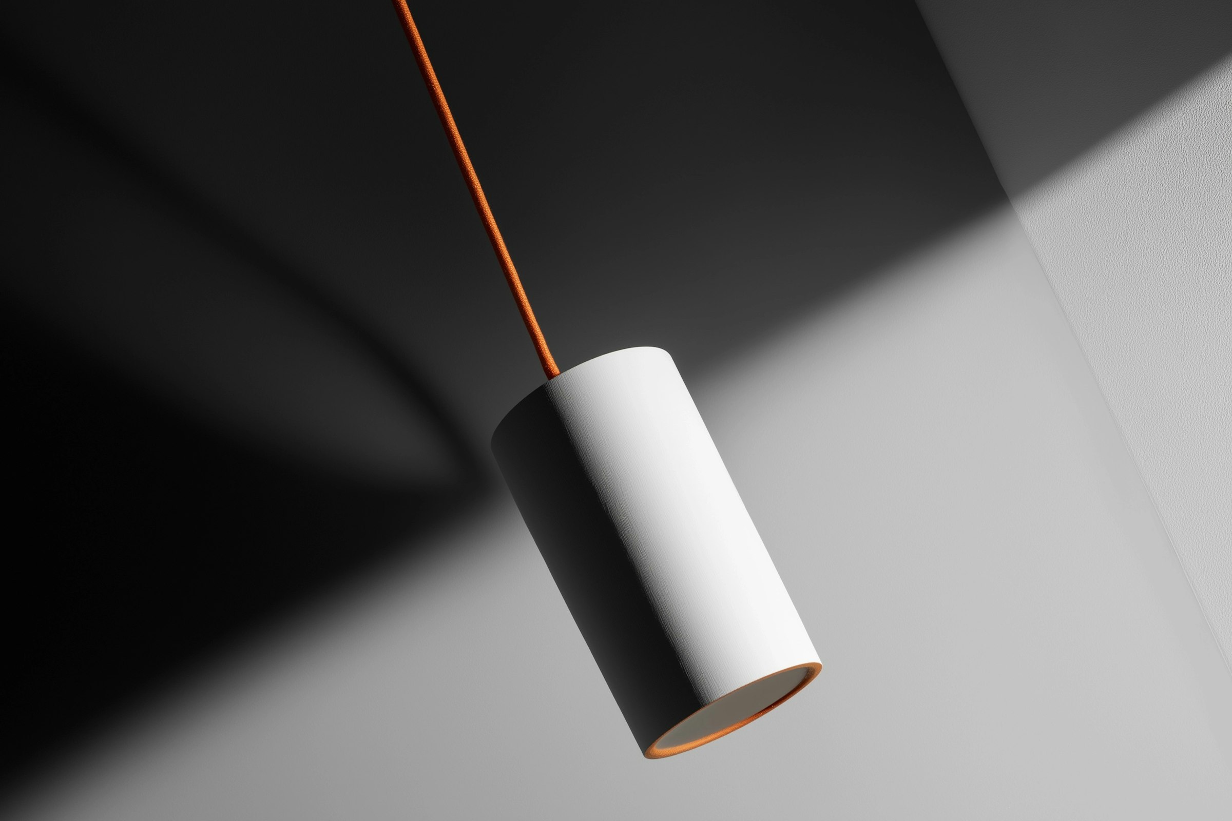Minimalist white cylindrical pendant light with orange cord against a dramatic light and shadow background.