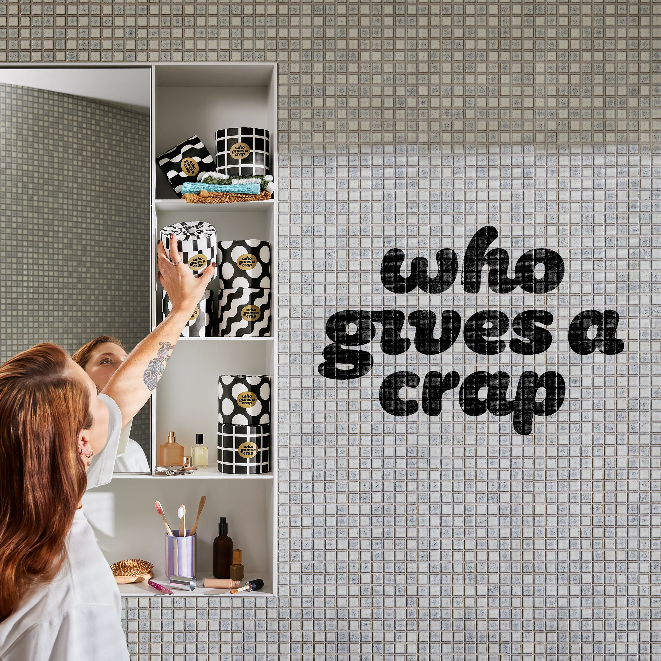 Person reaching into a bathroom shelf stacked with black-and-white patterned Who Gives A Crap rolls, the brand name across the tiled wall