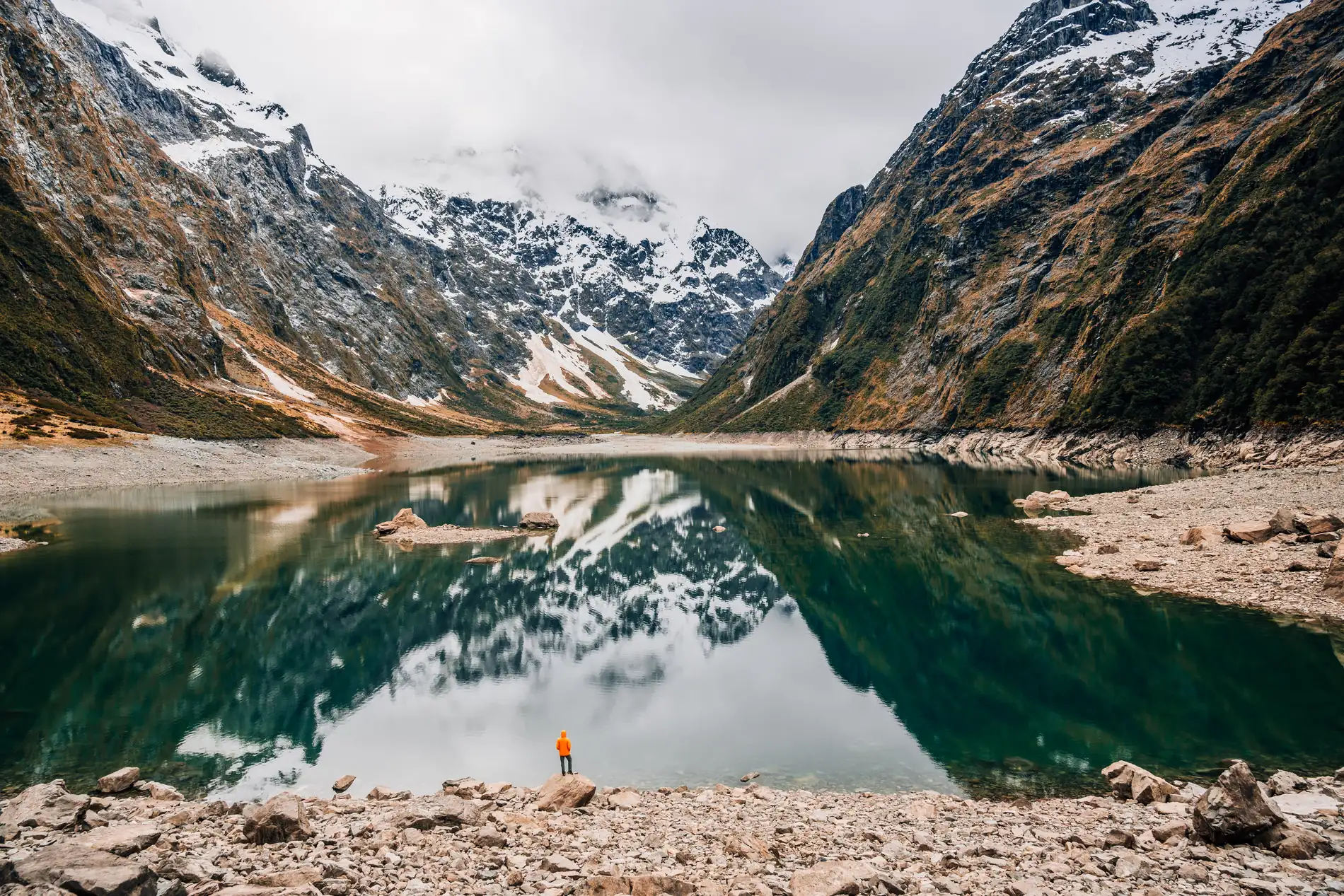 A glacial lake mirroring snow-streaked mountains, a lone figure on the stony shore