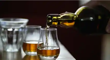 A bottle pours golden brown whiskey into a curved glass on a wooden table. A second glass and a blurred glass in the background add depth. Elegant and warm.